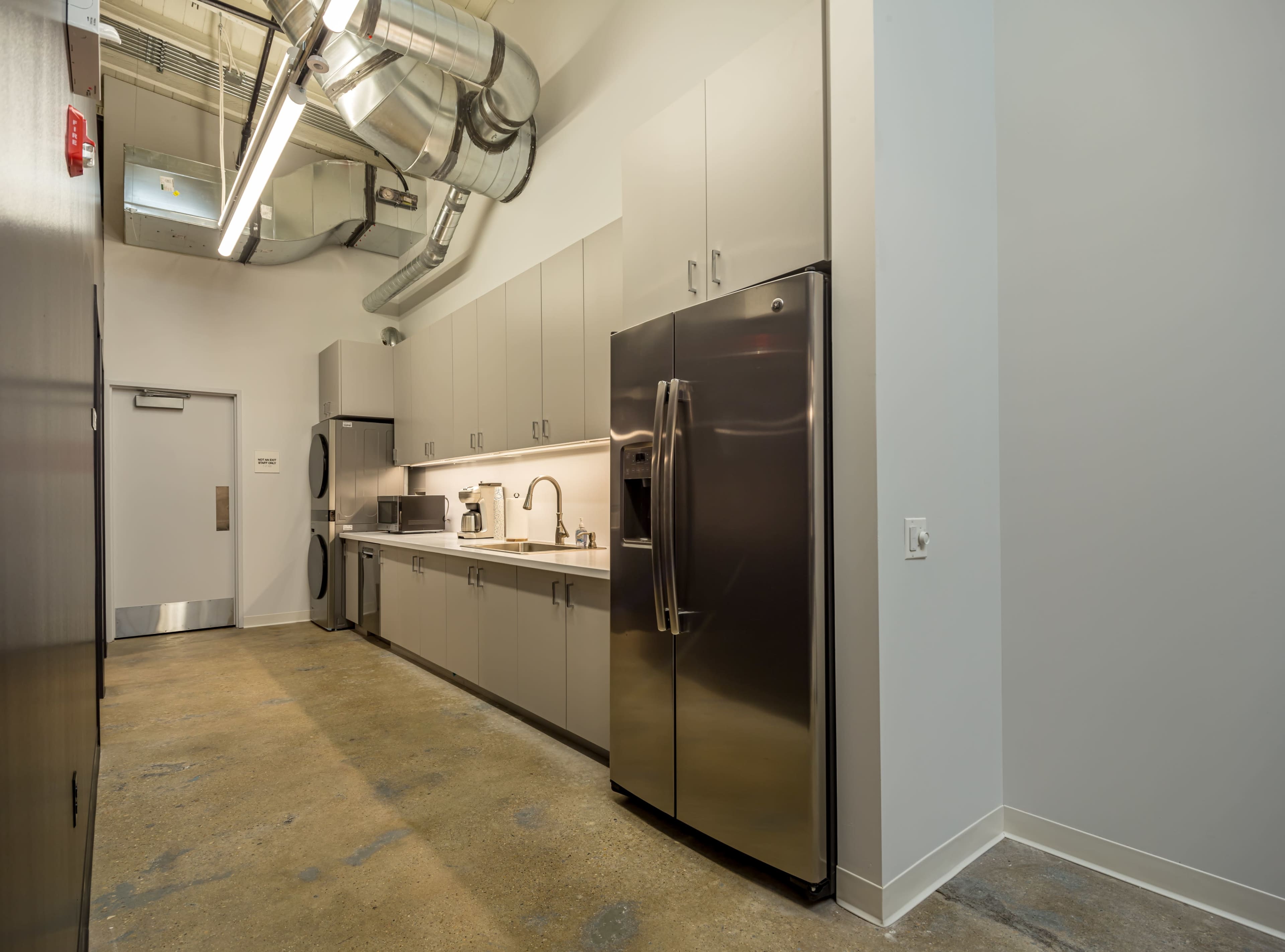The image shows a modern kitchen area with gray cabinets, stainless steel appliances, and a concrete floor.