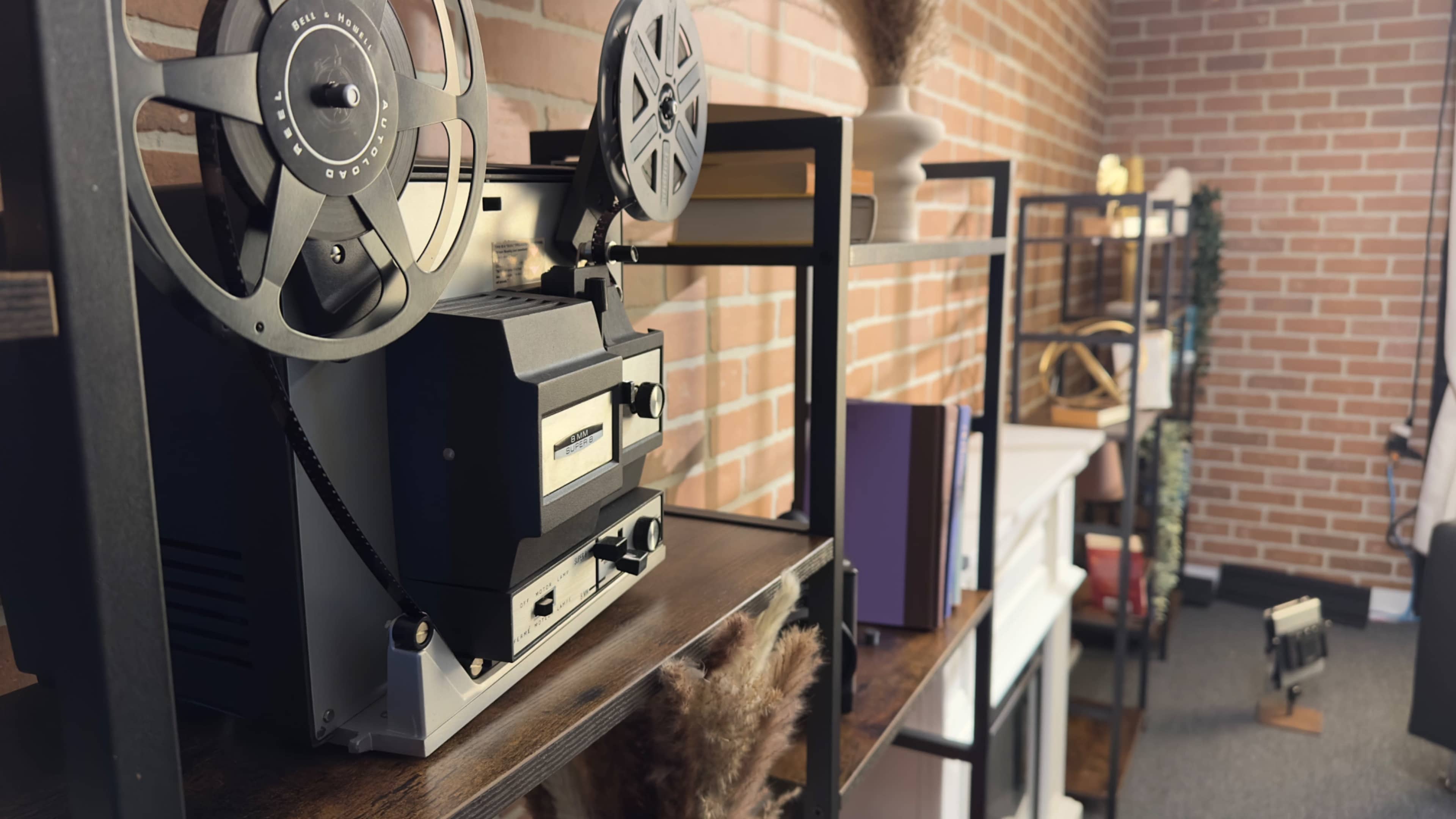 A vintage reel-to-reel tape recorder is positioned on a wooden shelf in a room with exposed brick walls and various decorative items.