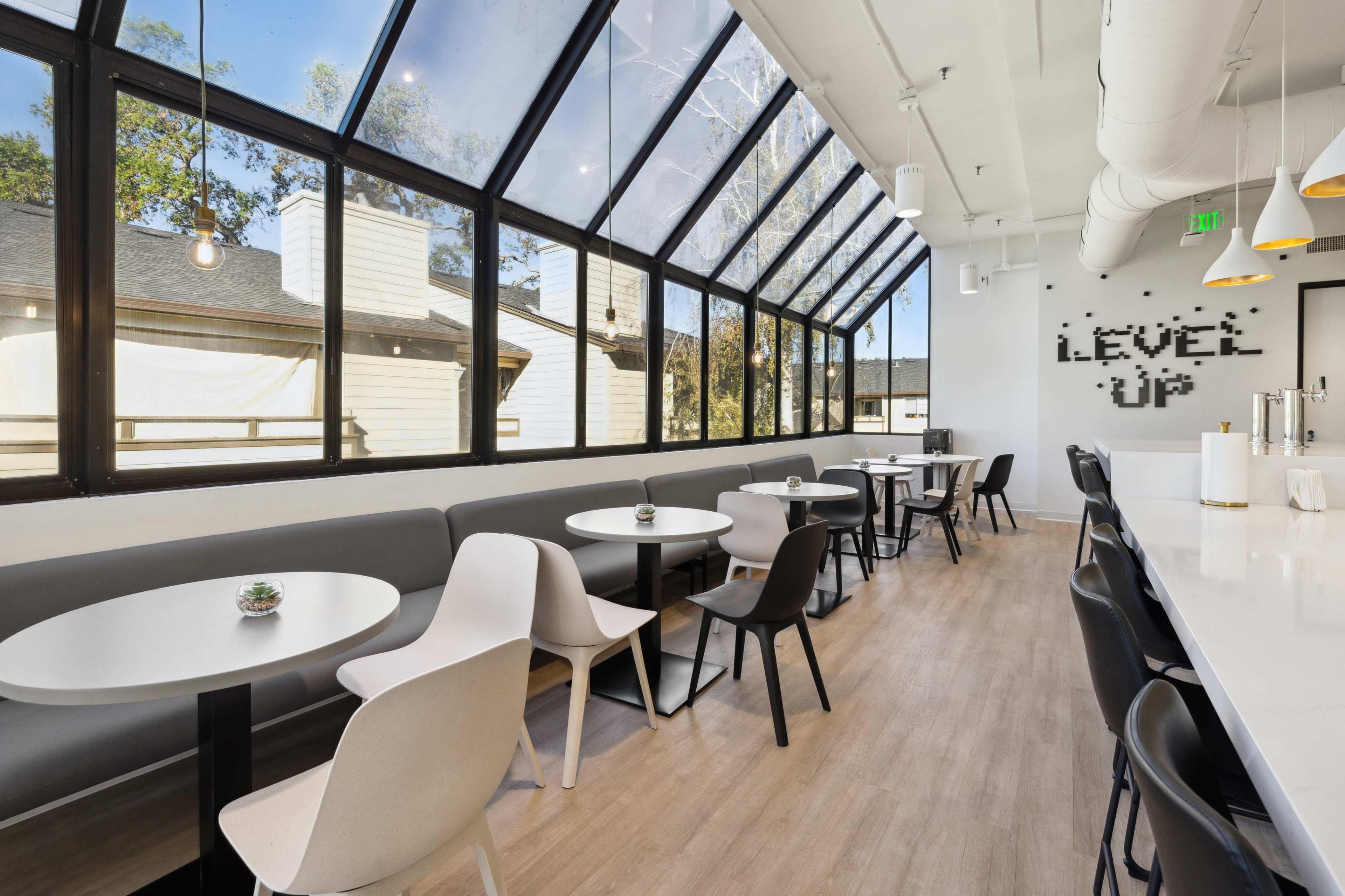 The image shows a modern café interior with large windows, white tables, and a mix of chairs arranged neatly along a light wood floor.