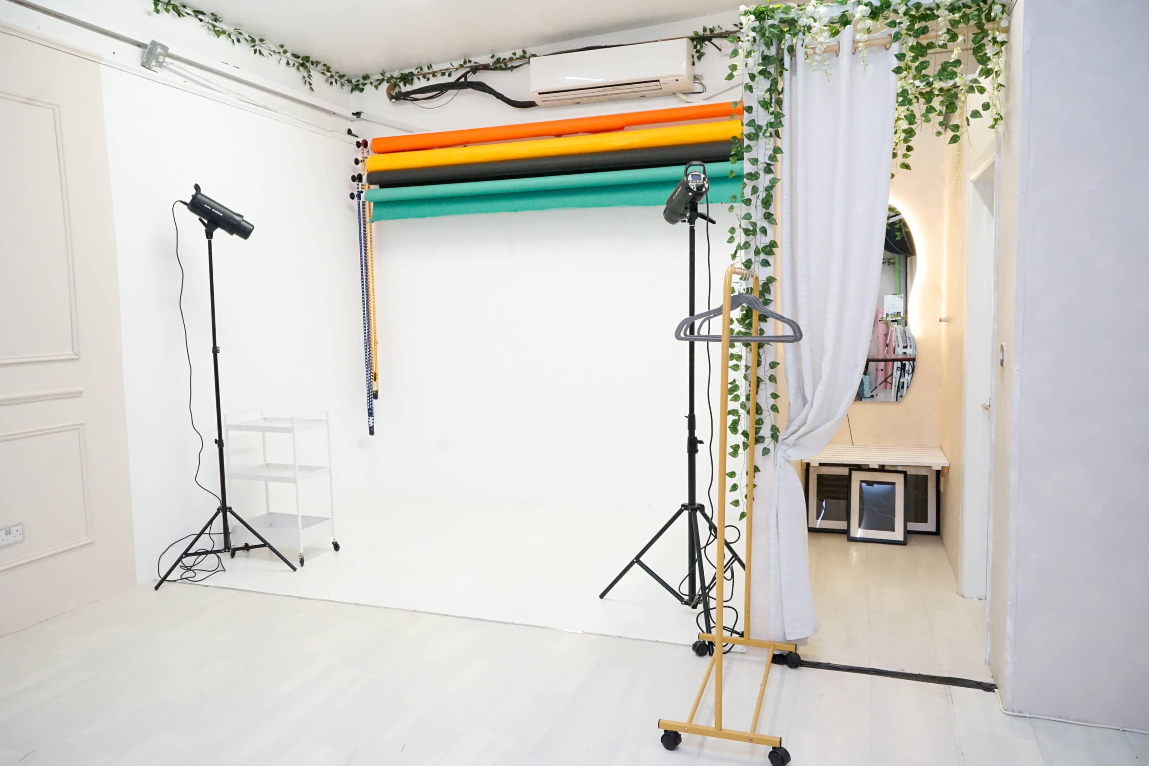 The image shows a photography studio with a white backdrop, colorful photography rolls, and lighting equipment, along with a clothing rack and a mirror.