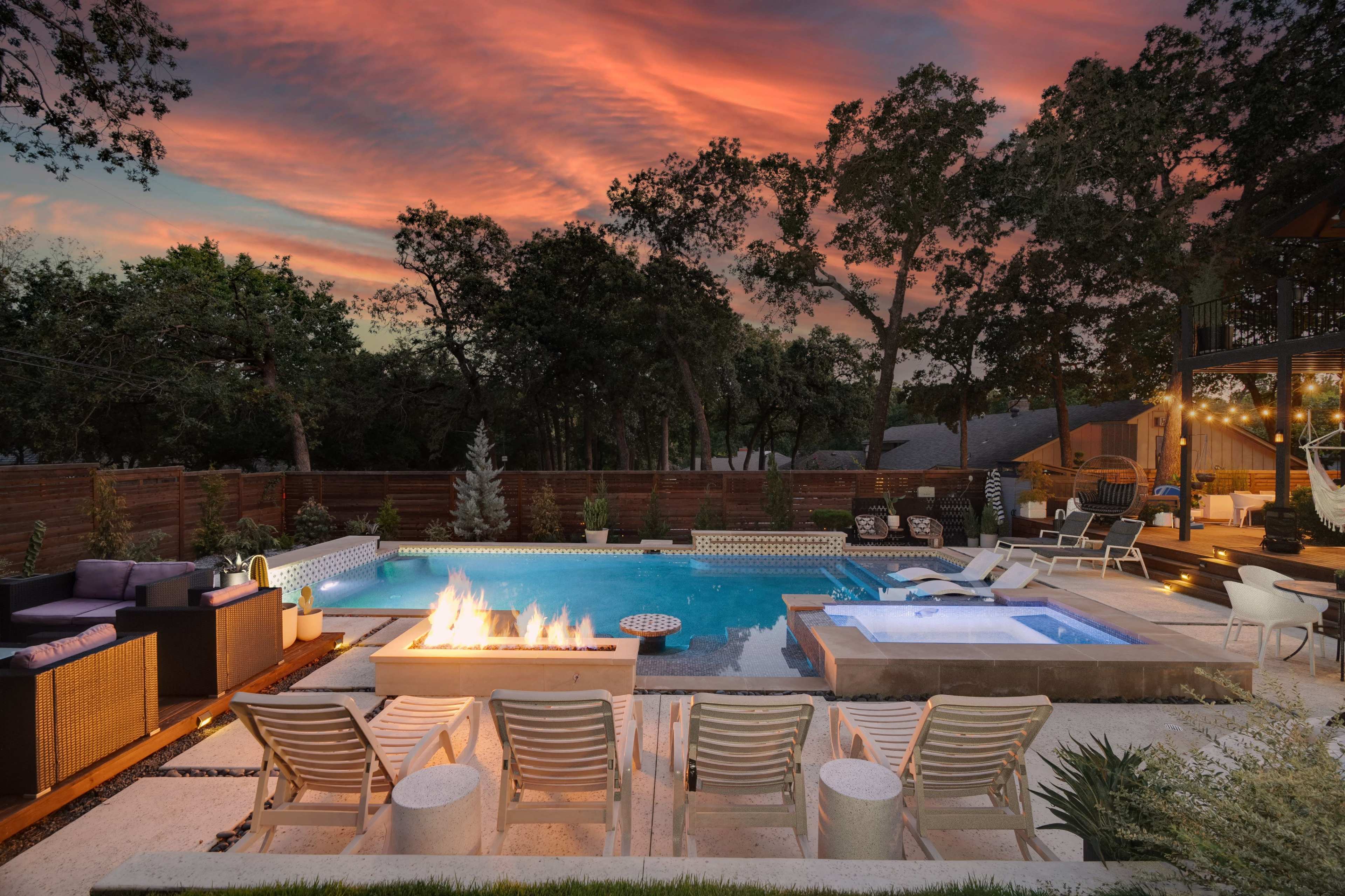 The scene features a stylish backyard pool area with lounge chairs, a fire pit, and a hot tub, surrounded by trees under a colorful sunset sky.