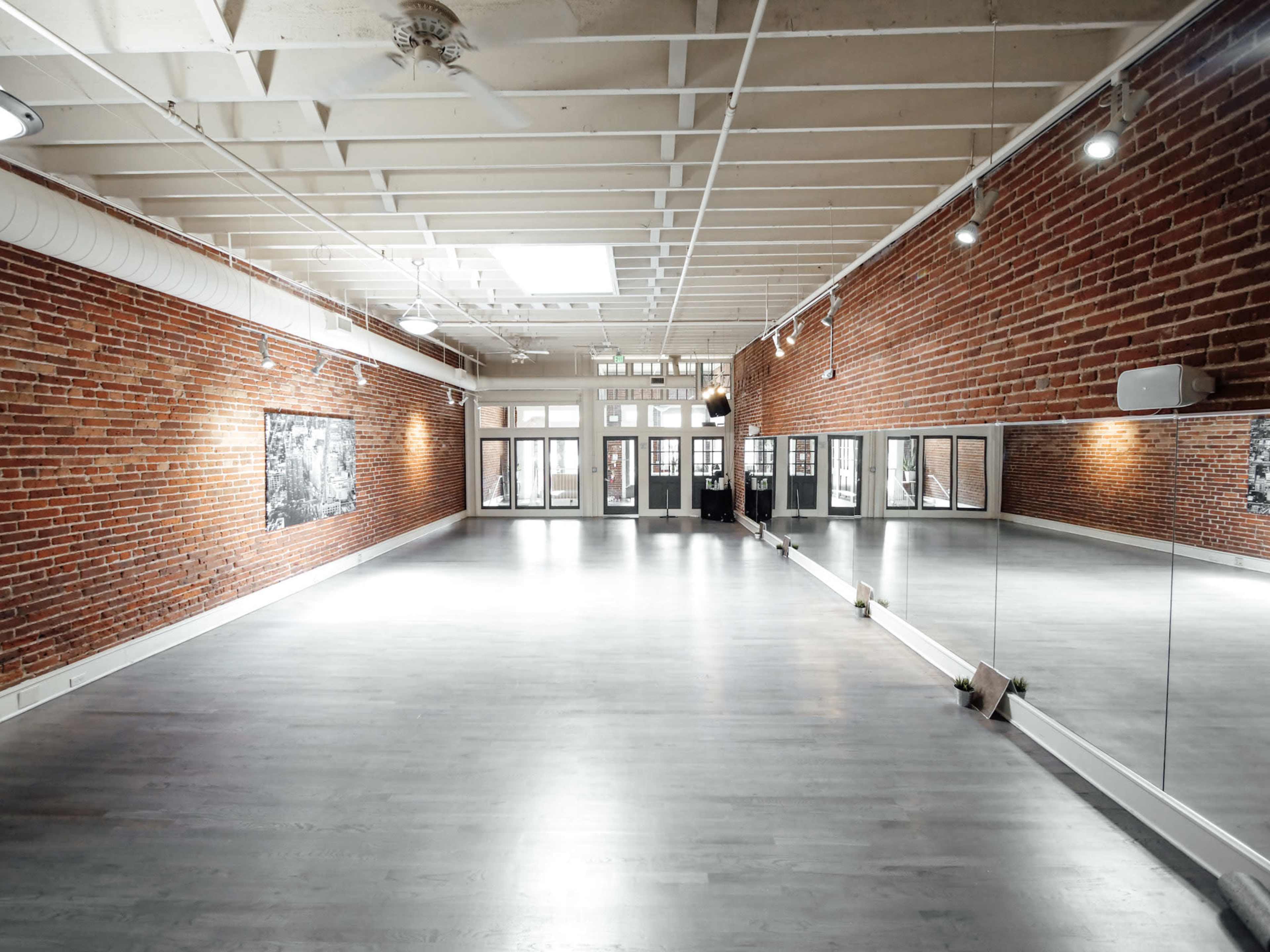 The image shows a spacious, well-lit studio with exposed brick walls and a mirrored wall, featuring large windows that provide natural light.