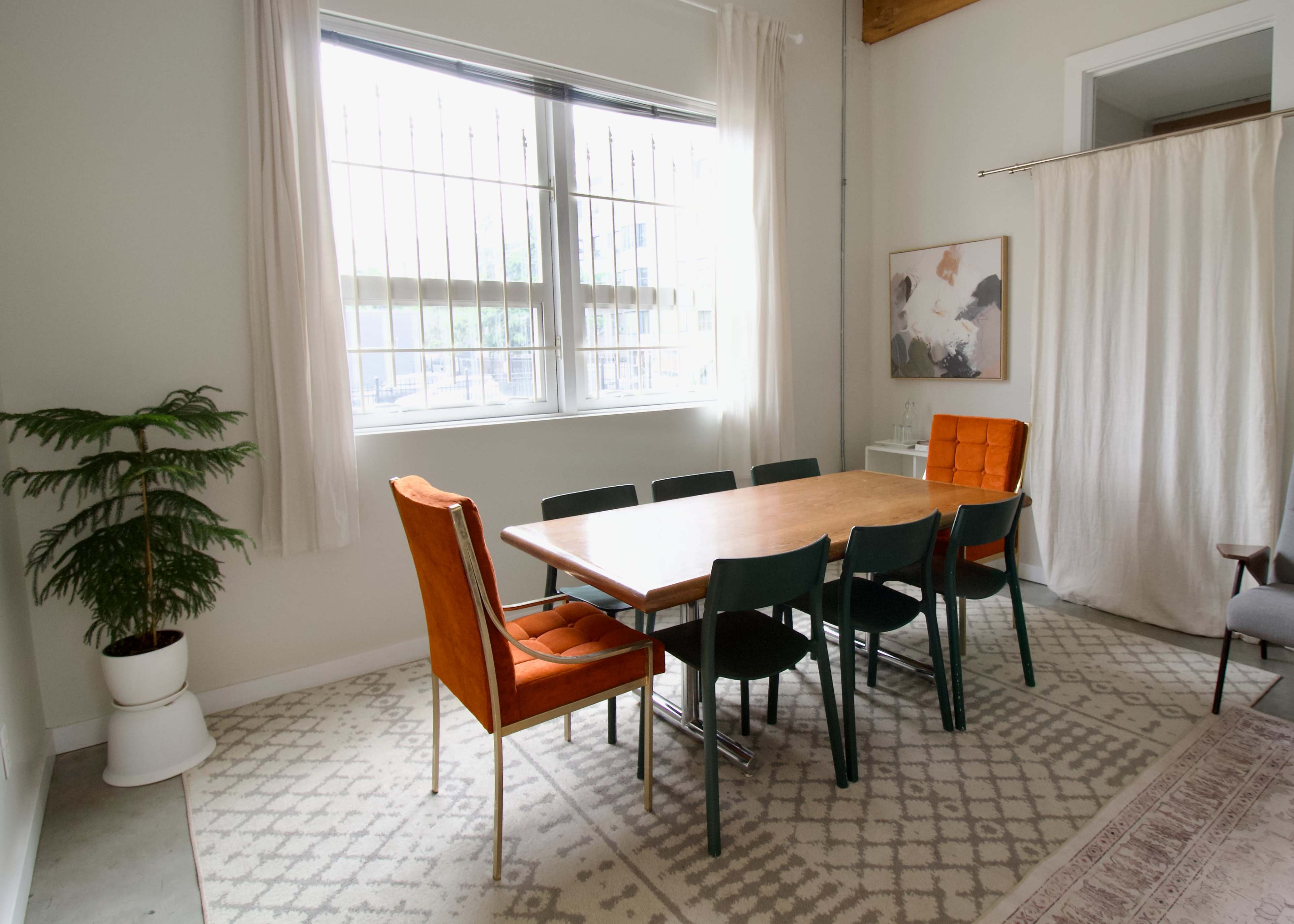 A dining area features a wooden table surrounded by eight chairs, with a large window and potted plant nearby.