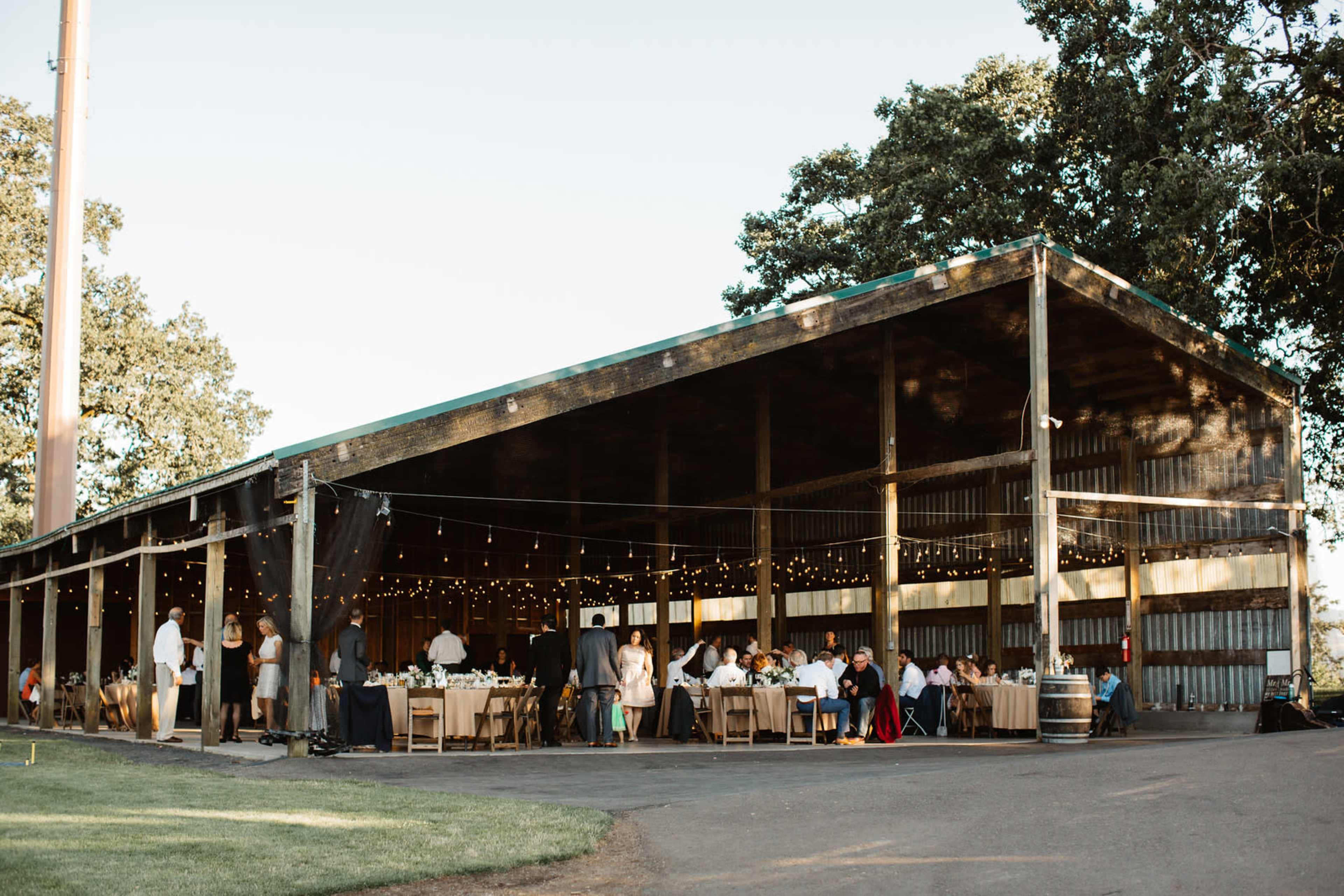 A large outdoor barn setting is filled with tables and guests celebrating, illuminated by string lights.
