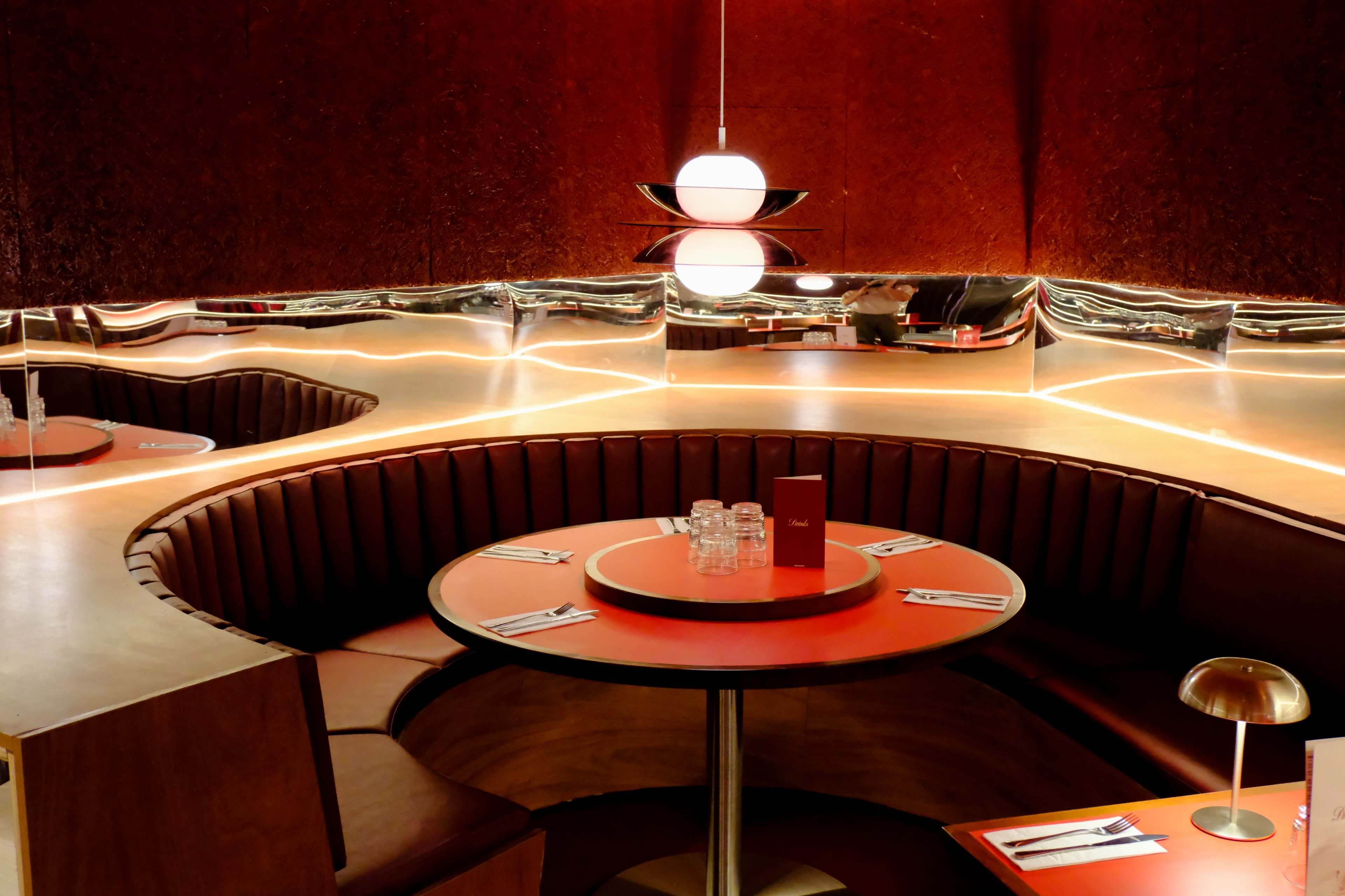 The image shows a modern restaurant interior featuring a circular booth with red upholstery and a reflective wall accent, illuminated by a pendant light.