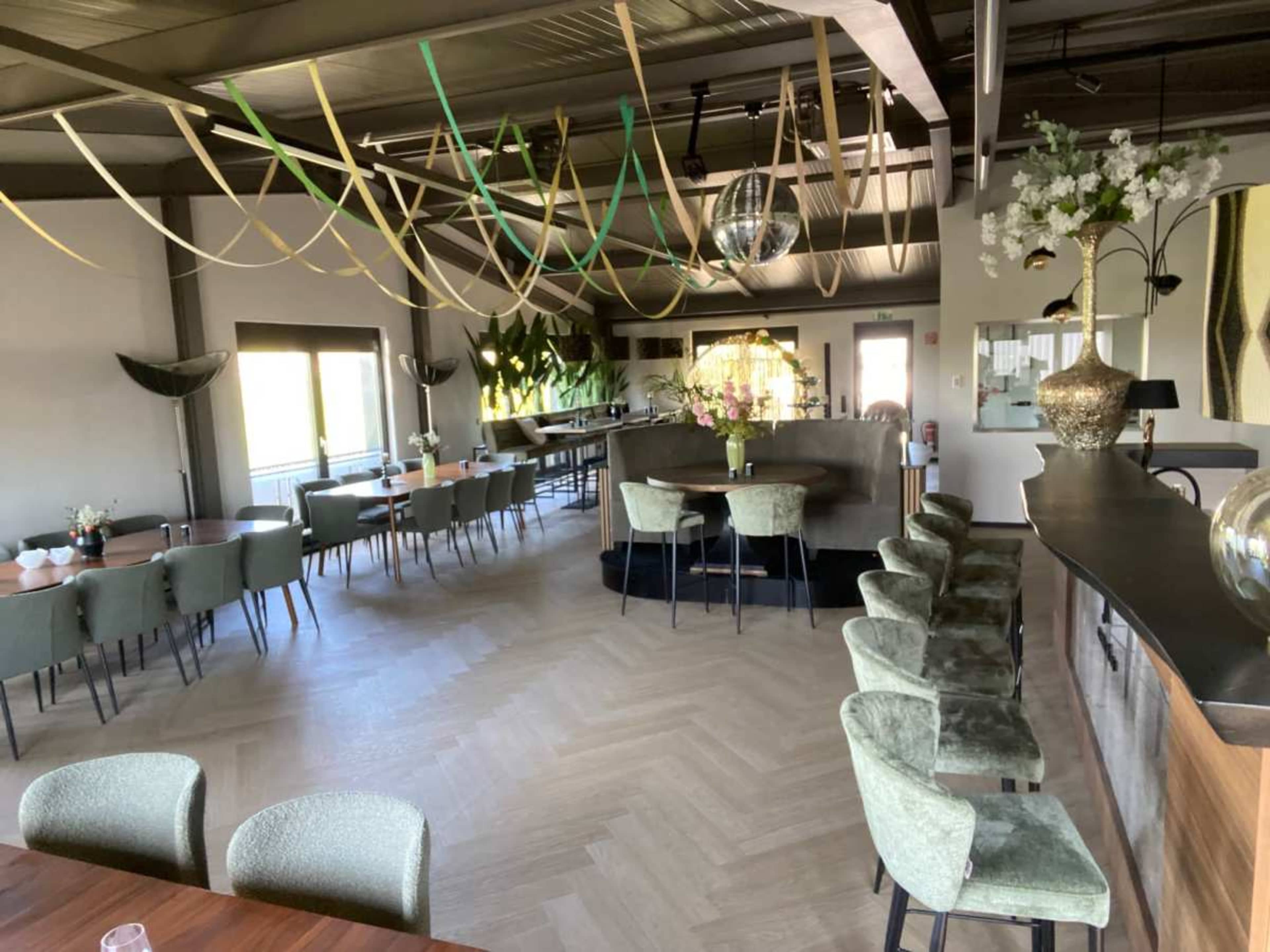 The image shows a modern dining area with tables and green seating, featuring decorative plants and hanging colored ribbons from the ceiling.