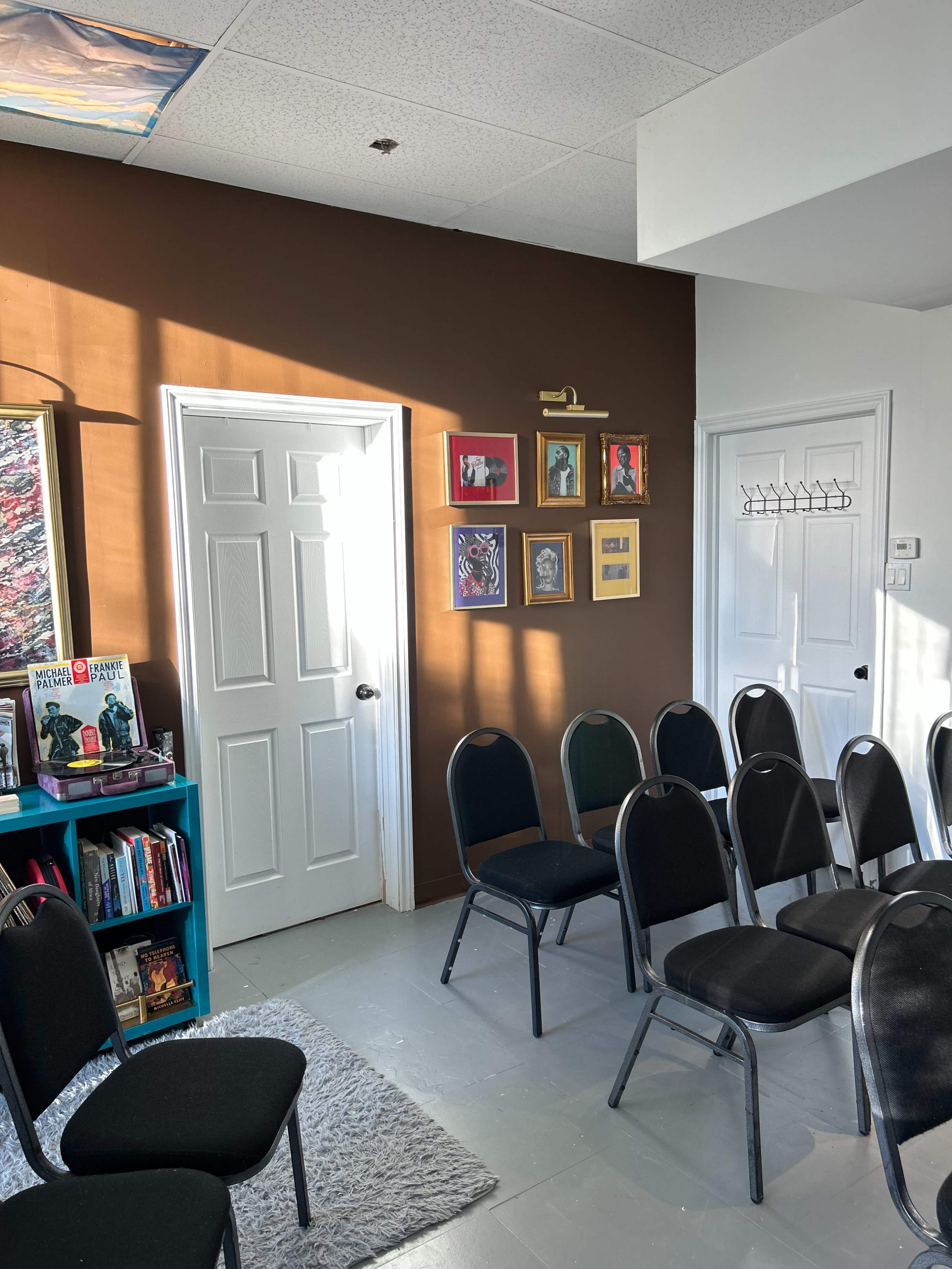 The image shows a room with a brown wall, a door, framed artwork, a bookshelf, and several black chairs arranged in rows.