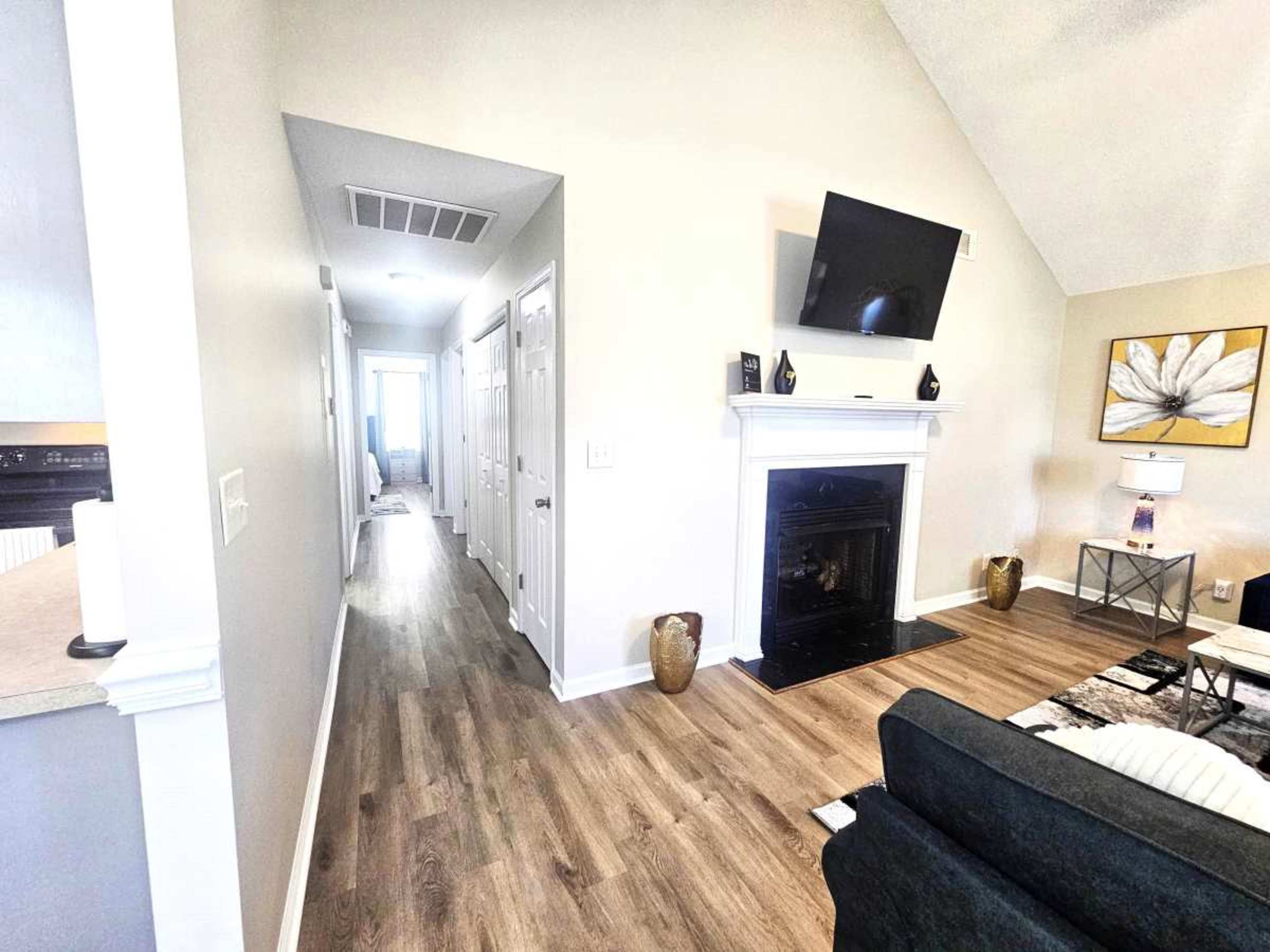 The image shows a well-lit living space featuring a fireplace, a mounted television, and a hallway leading to other rooms.