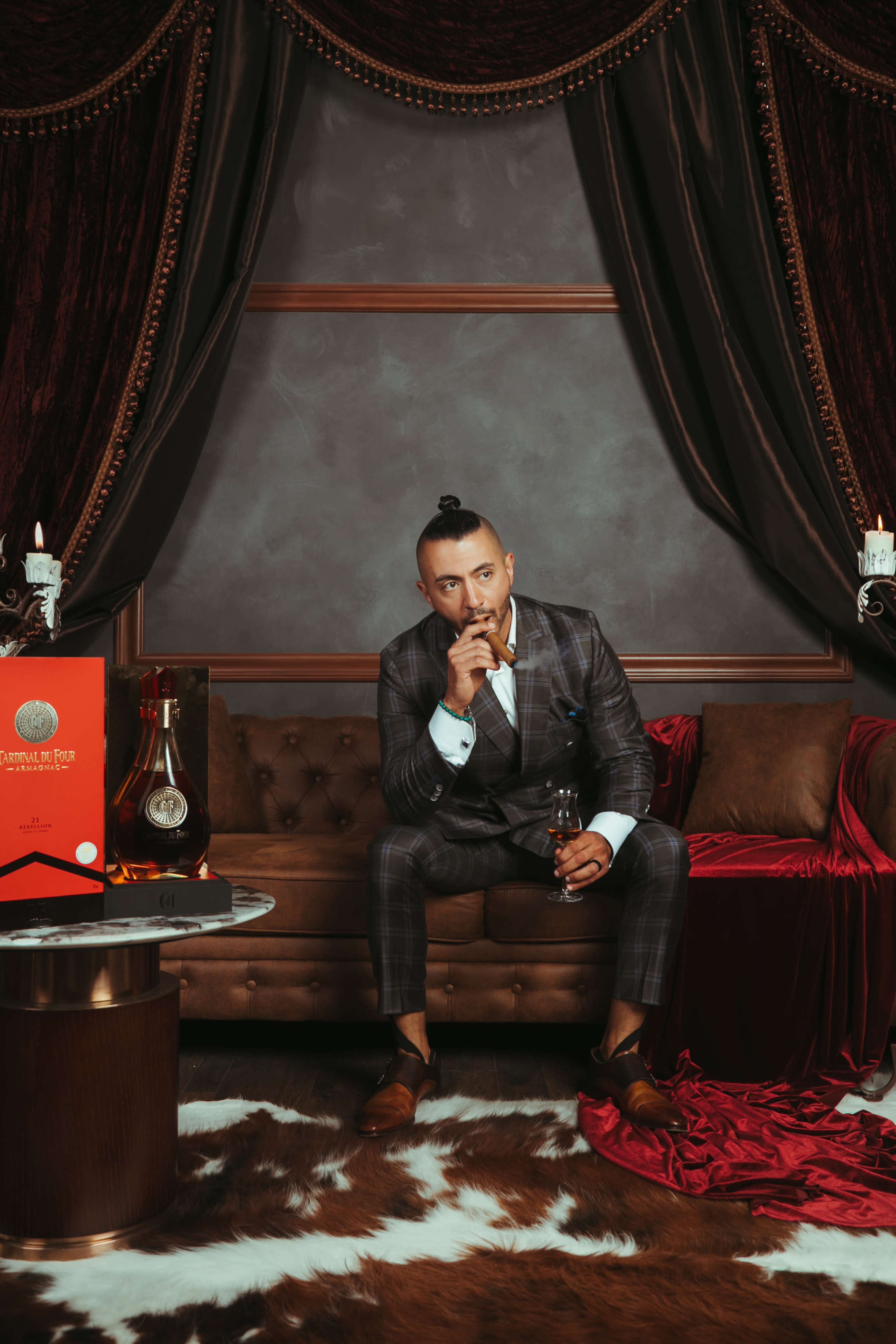 A man in a plaid suit sits on a brown velvet sofa, holding a drink, with luxurious decor and a bottle of liquor nearby.