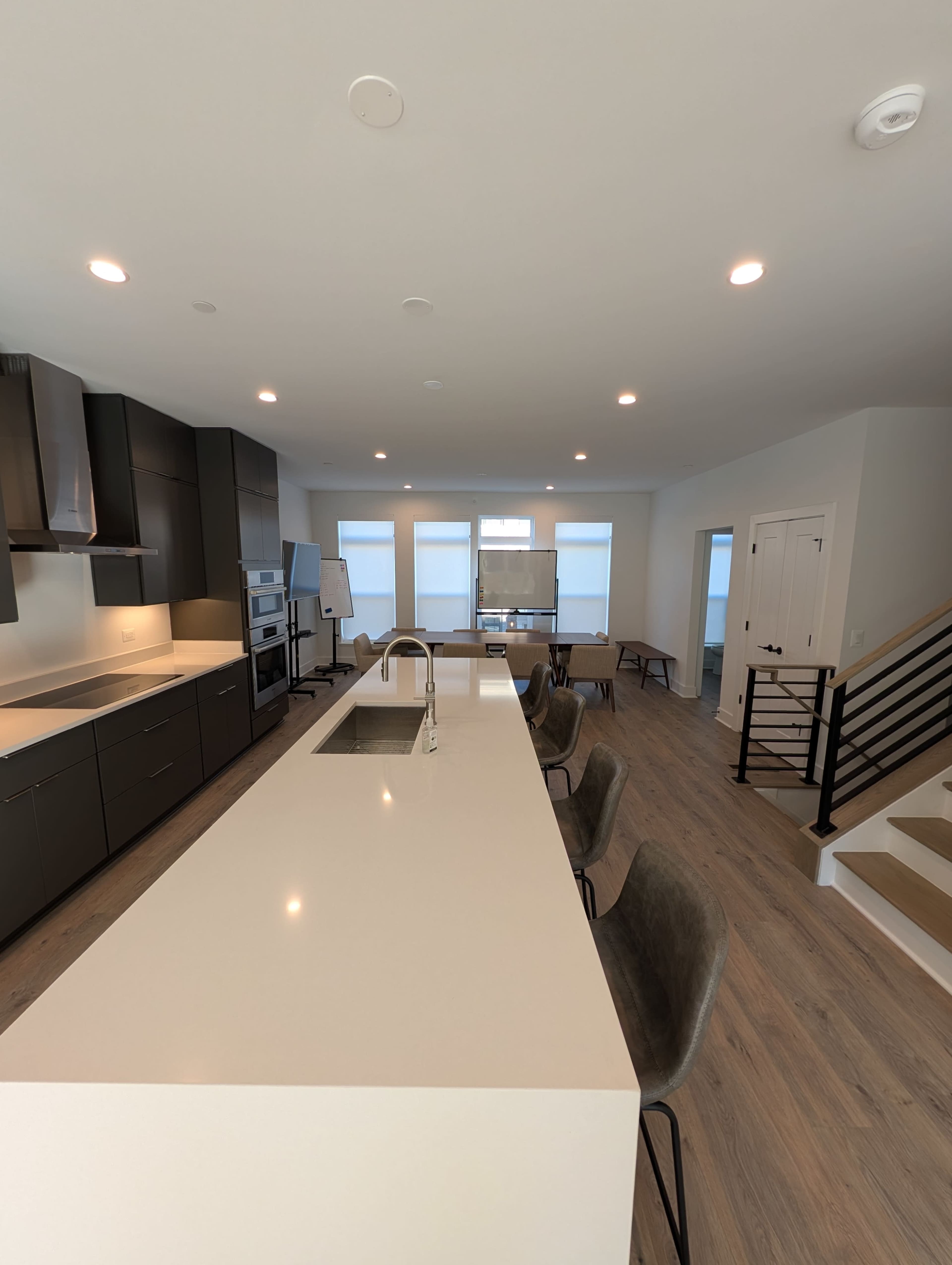 The image shows a modern kitchen with a large white island, dark cabinetry, and a spacious living area featuring large windows.