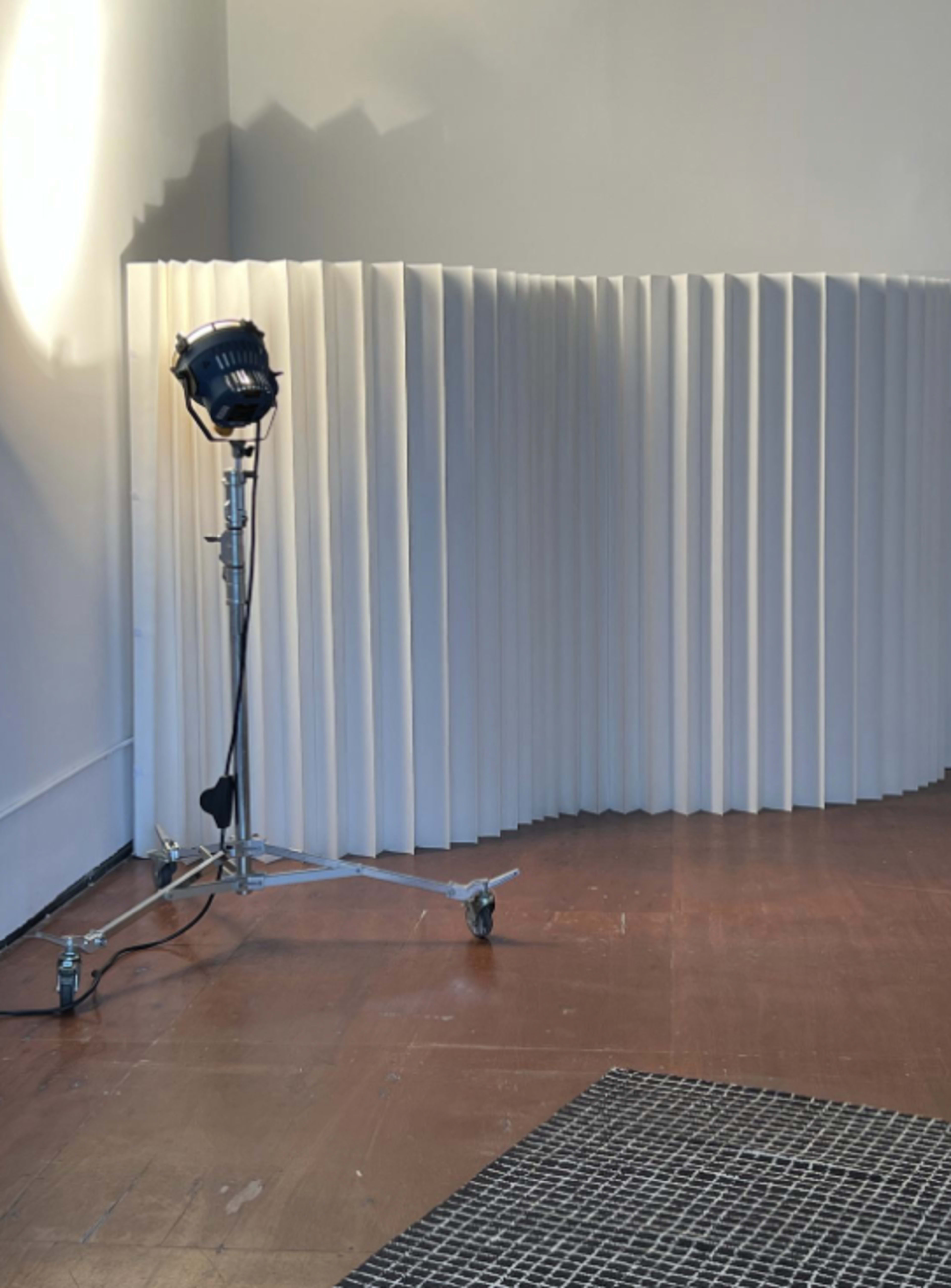 A floor-mounted spotlight casts light on a curved, textured white partition in a minimalist room with hardwood flooring.
