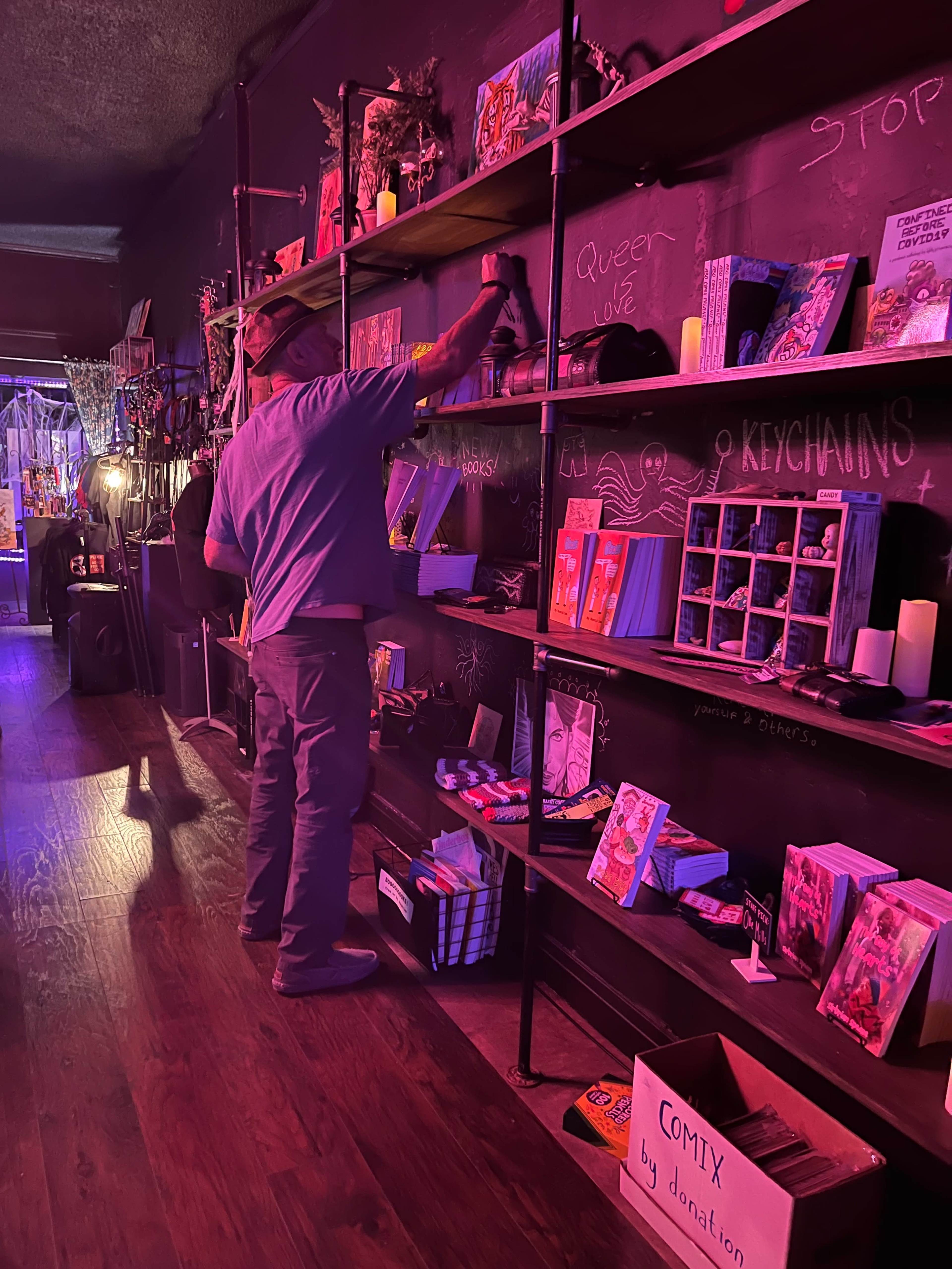 A person in a blue shirt is organizing books on a shelf in a dimly lit space lined with various displays and items.