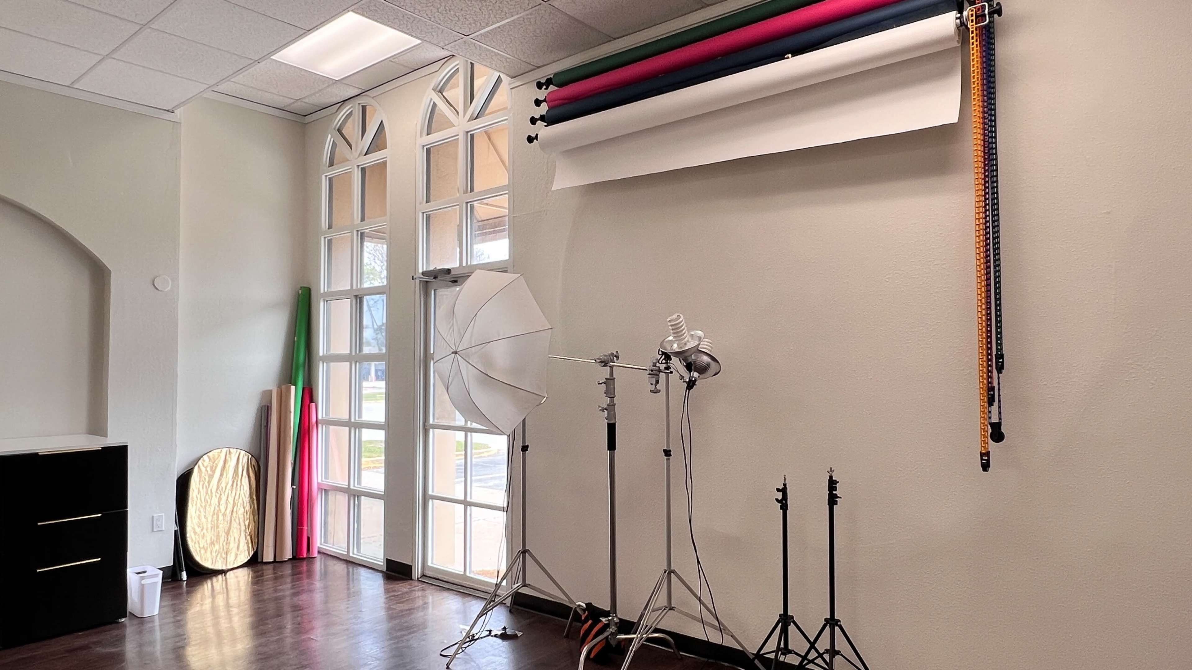 A photography studio with background rolls, lighting equipment on stands, and a large reflector against a neutral wall.
