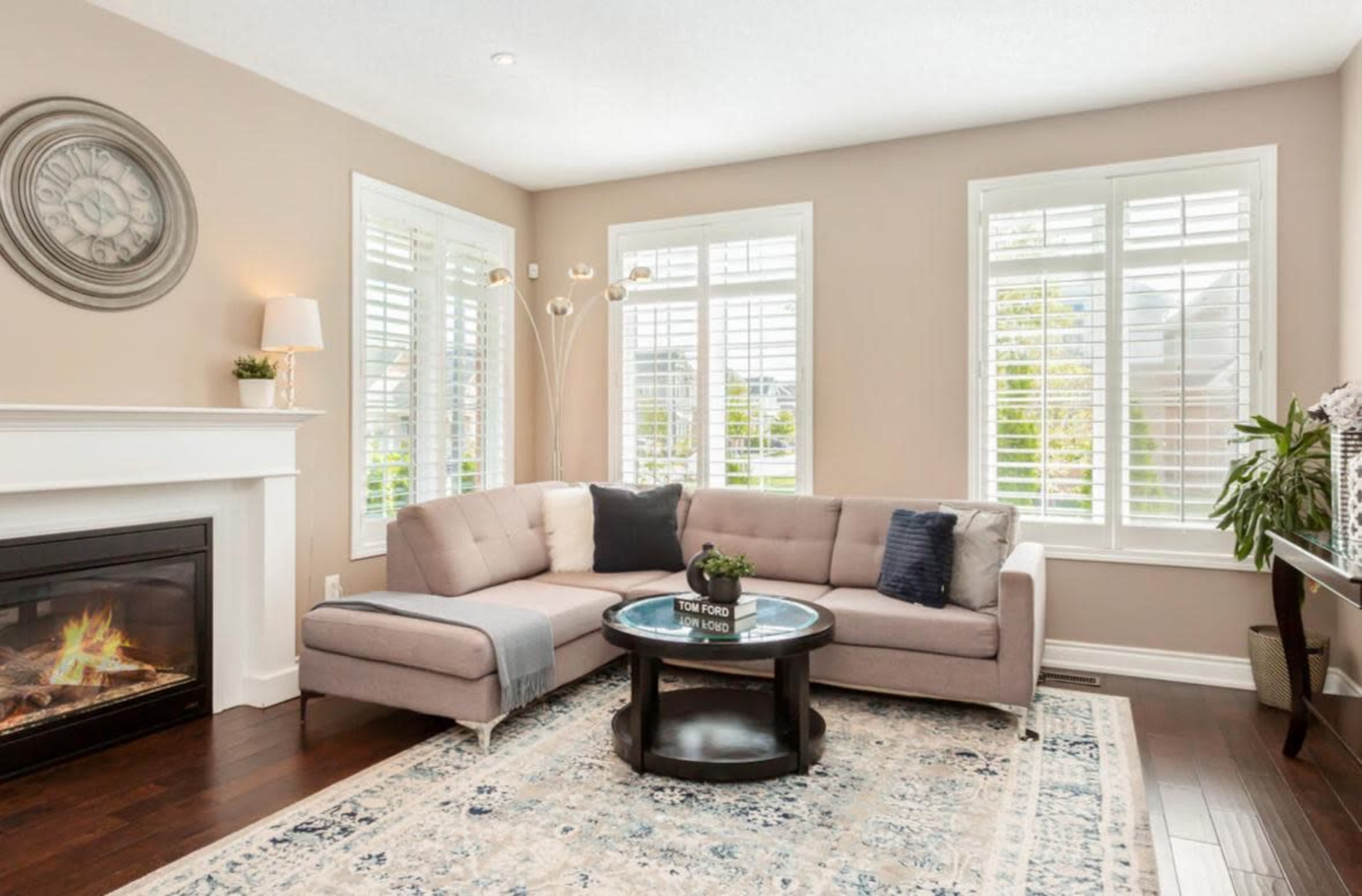The image shows a modern living room with a sectional sofa, a round coffee table, and large windows with plantation shutters.