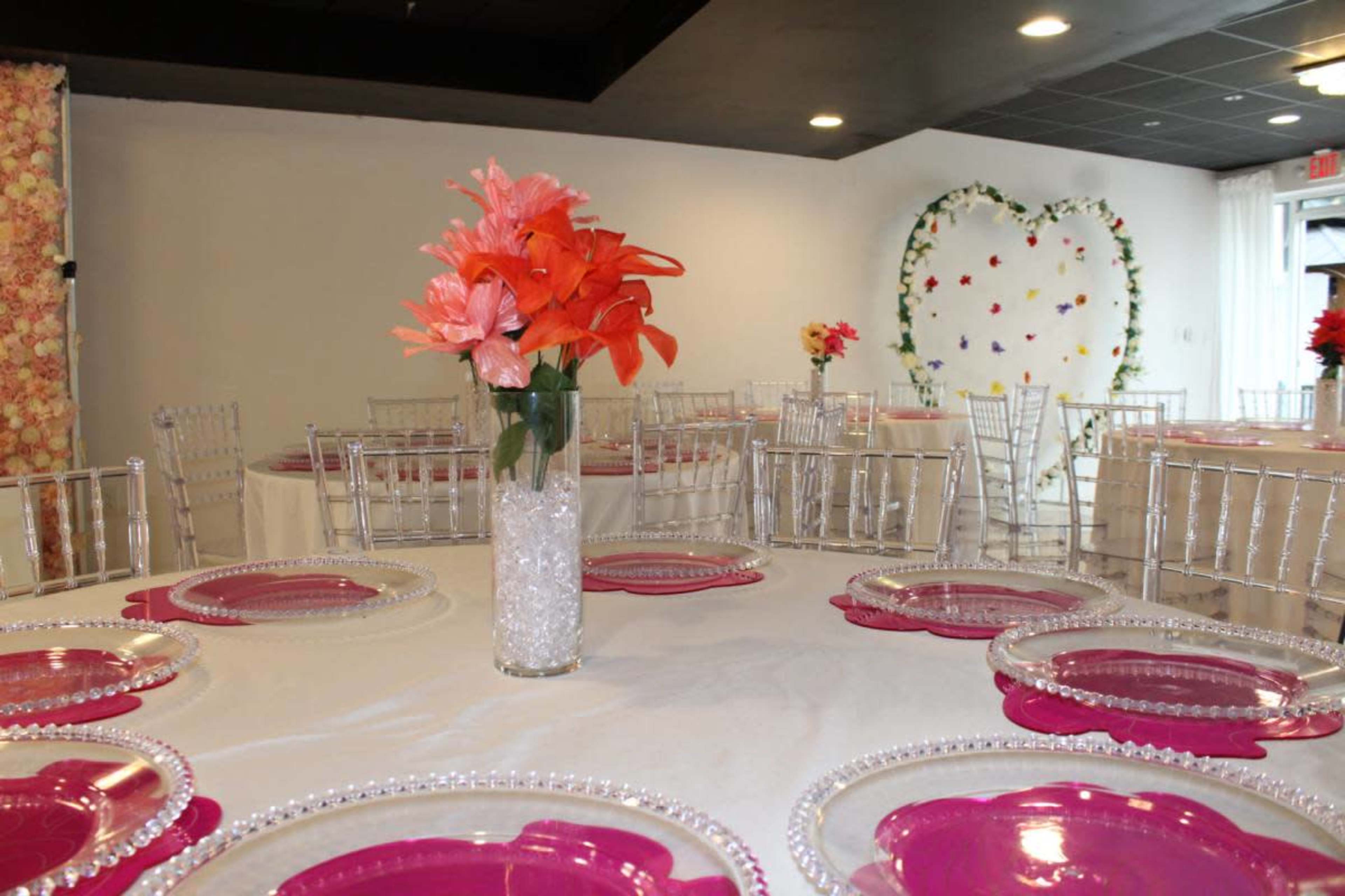 The image shows a banquet hall set with round tables adorned with pink placemats and clear plates, featuring floral centerpieces in glass vases.