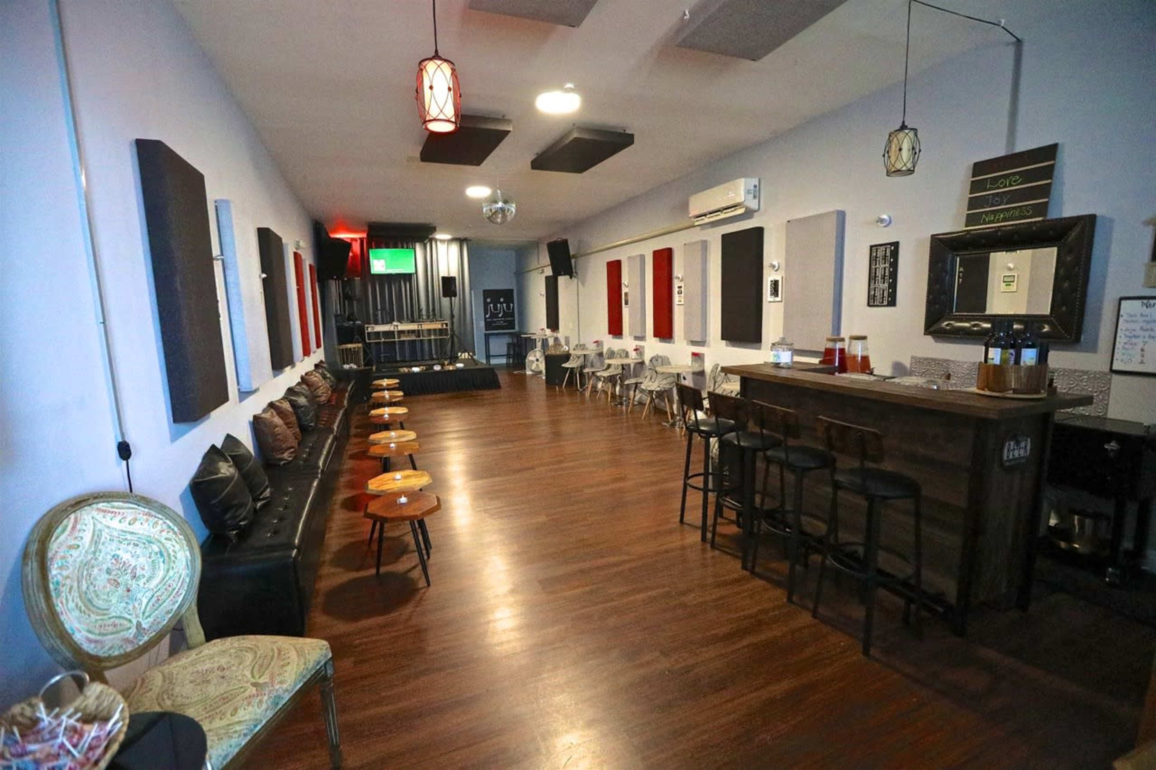 A spacious lounge with a bar, wooden flooring, and chairs arranged around small tables, along with acoustic panels on the walls.