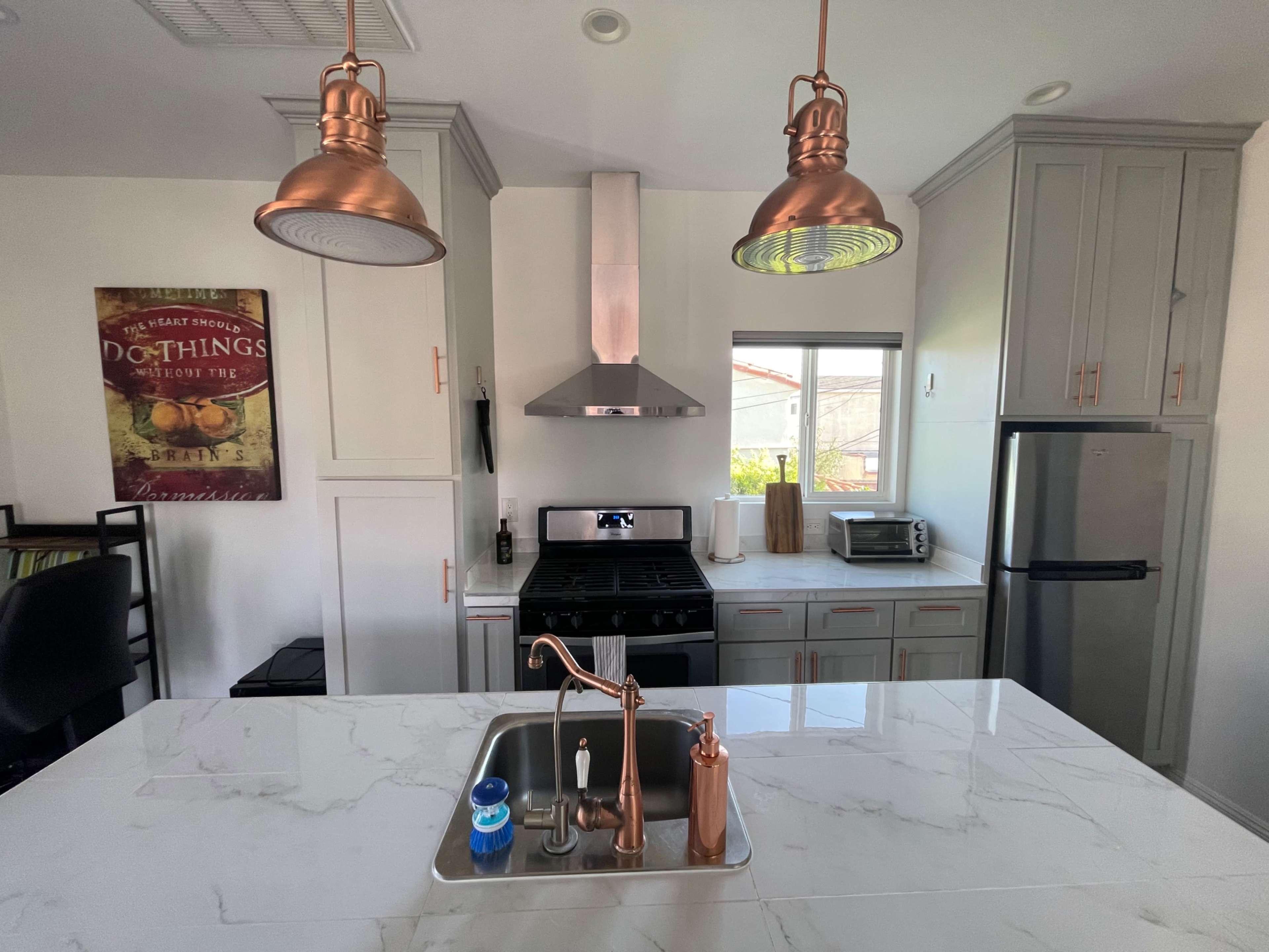 The image shows a modern kitchen featuring copper pendant lights, a stainless steel range hood, and a marble countertop with a sink.