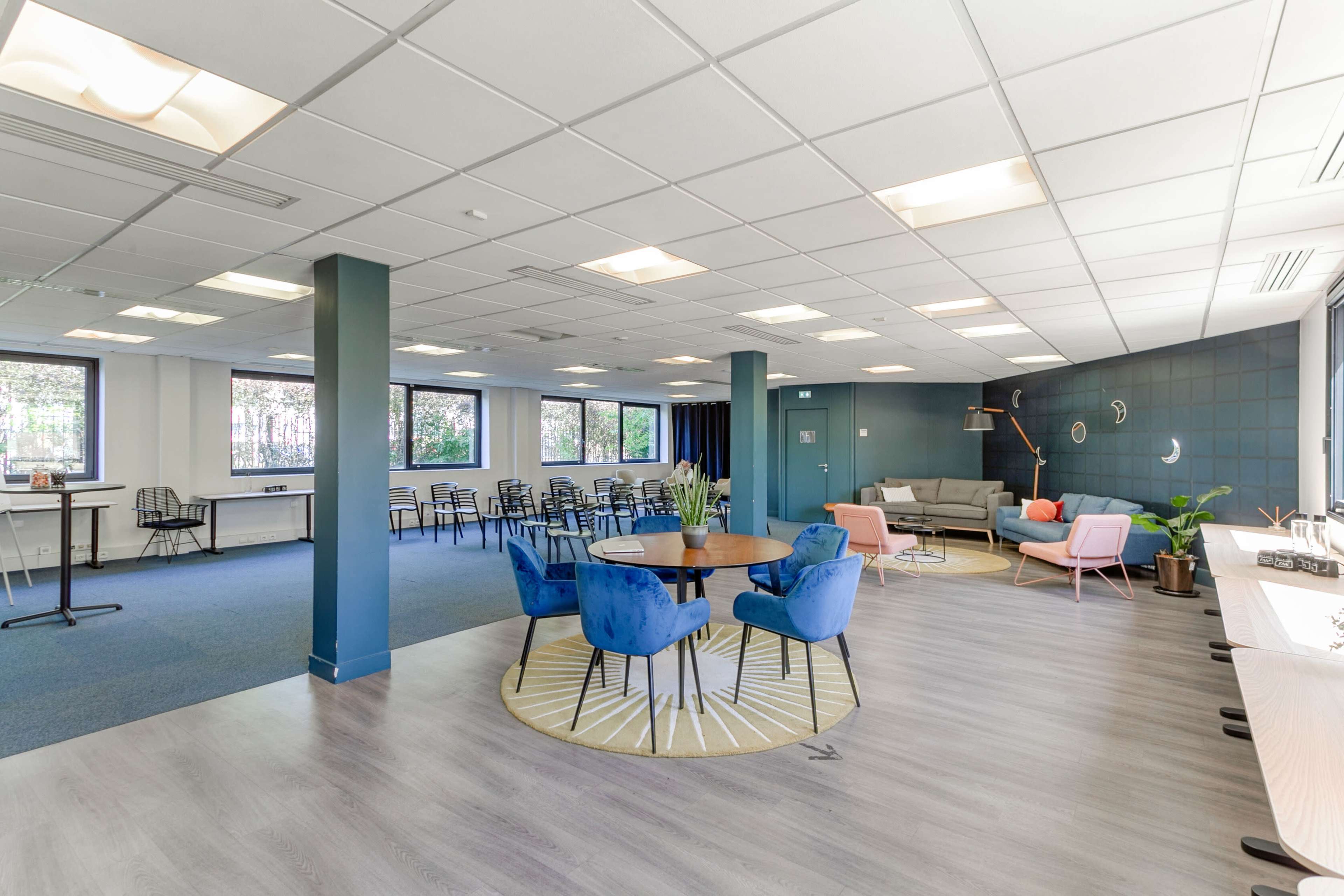 A modern office space with a mix of seating arrangements, including blue chairs and a round table, alongside a lounge area with a sofa and plants.
