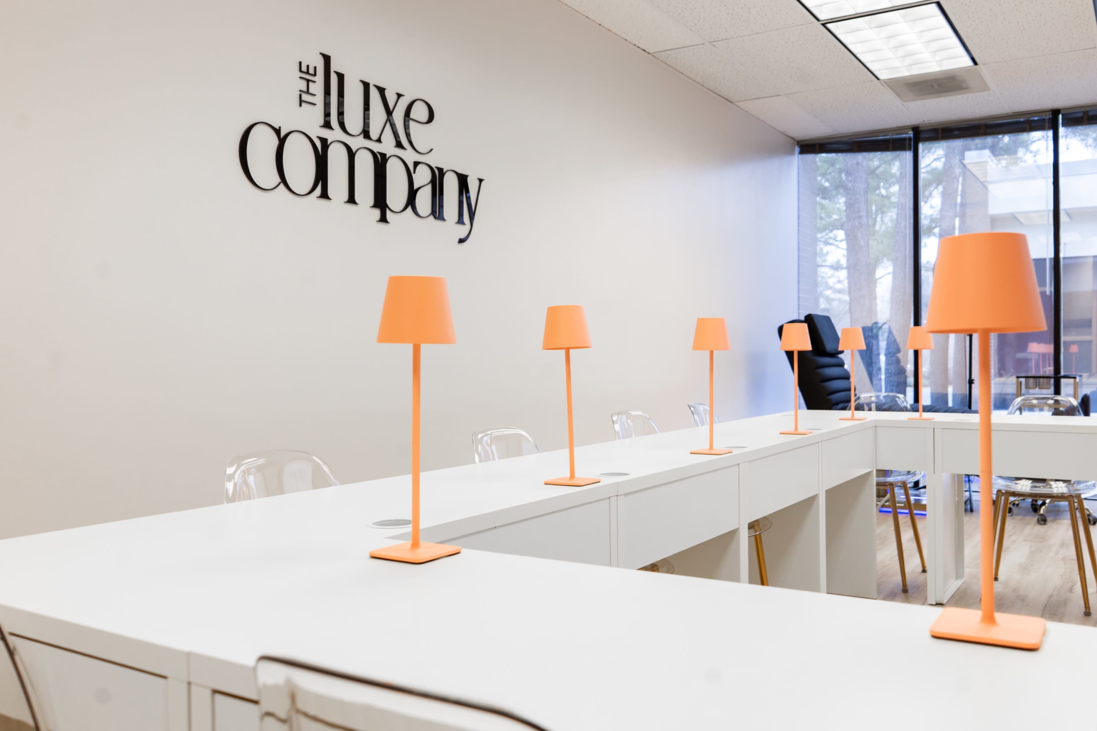 A modern conference room features a long white table with orange lamps and a prominently displayed sign reading "The Luxe Company" on the wall.