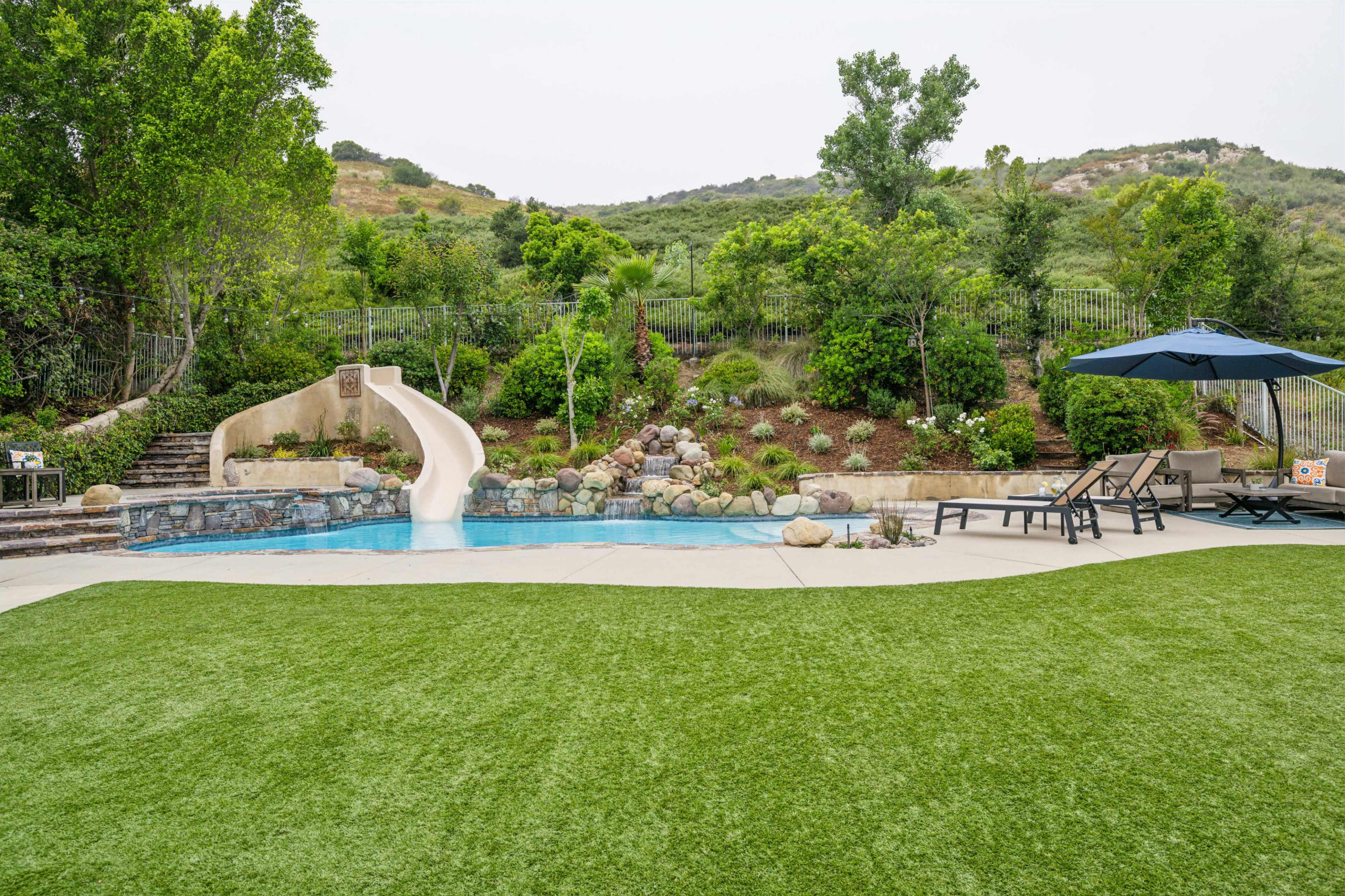 A backyard featuring a swimming pool with a slide, surrounded by landscaped greenery and a seating area under an umbrella.