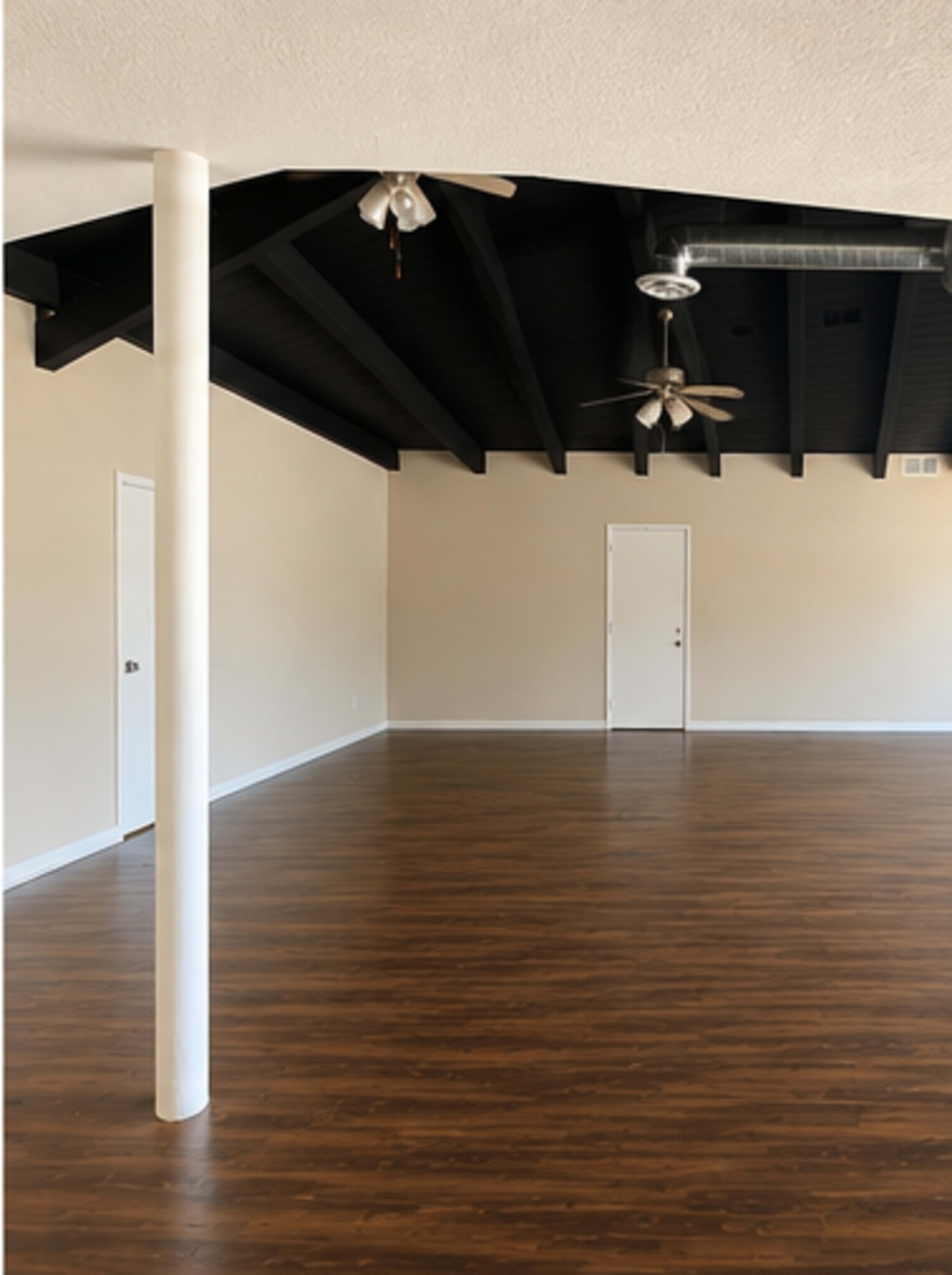 The image shows a spacious room with a polished wooden floor, a black-beamed ceiling, and a white column in the center.