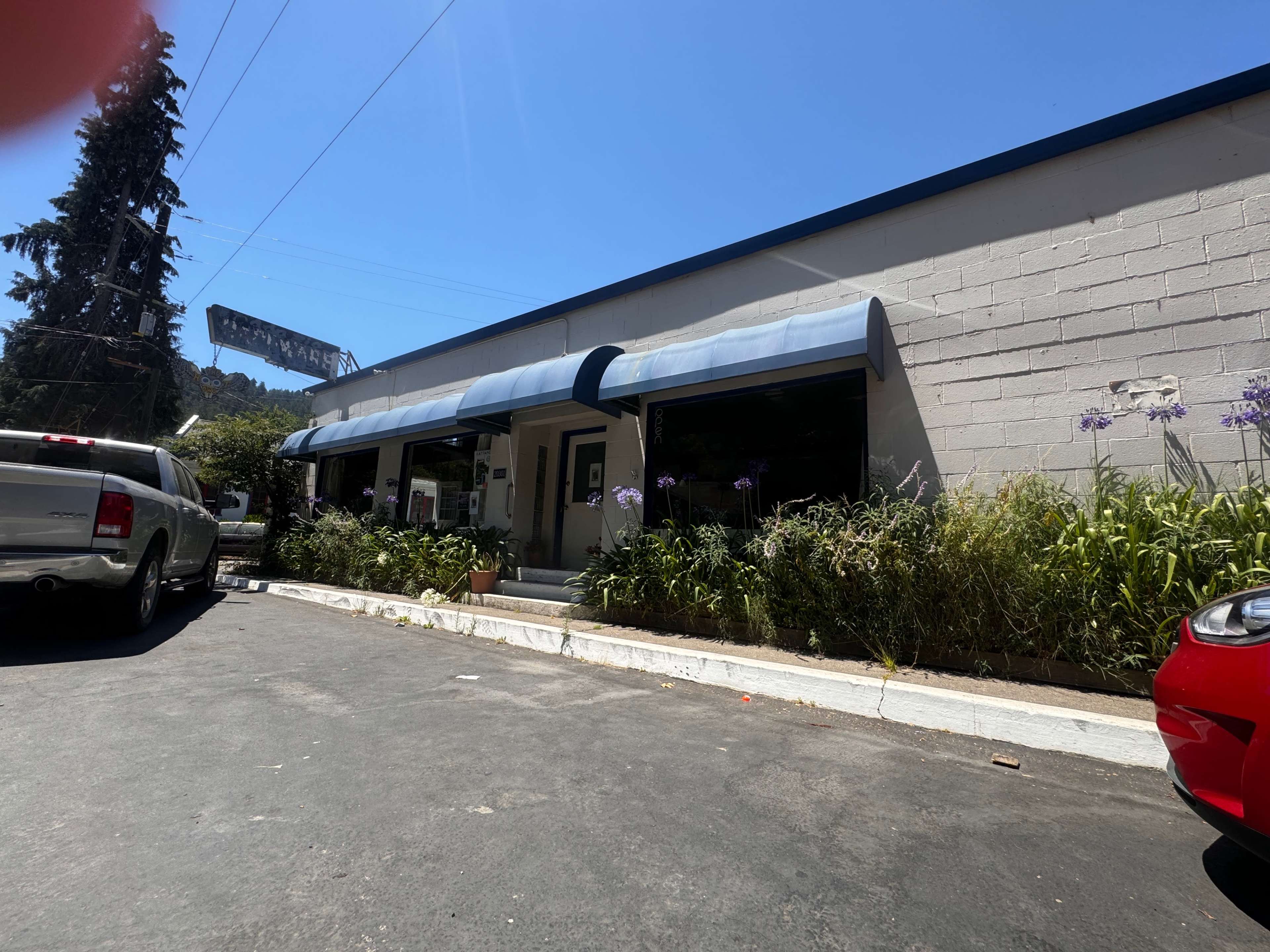 A single-story commercial building with an awning, surrounded by greenery, located on a paved driveway next to parked vehicles.
