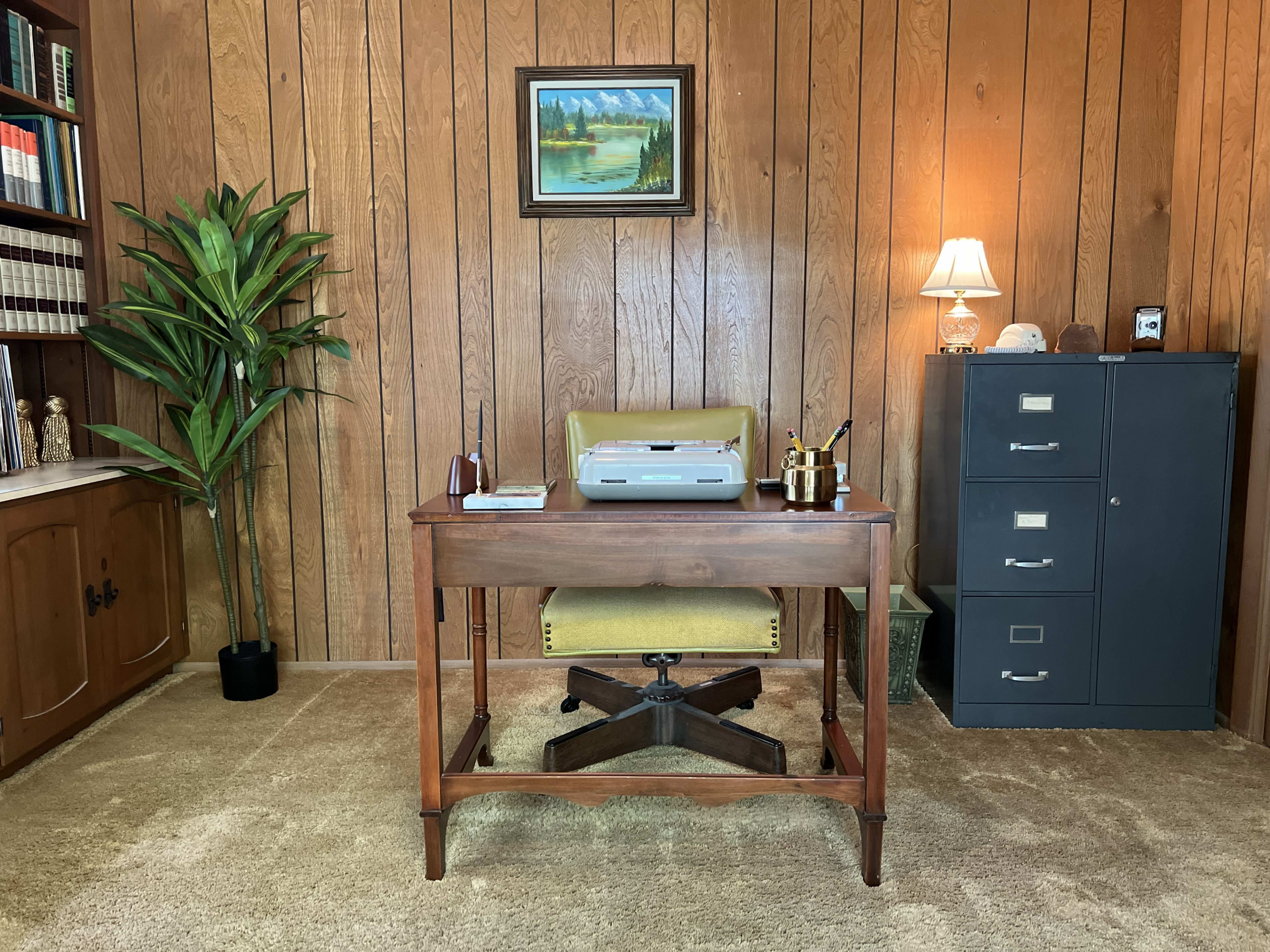 The image shows a home office with a wooden desk, a printer, a chair, a bookcase, a filing cabinet, and decorative items against a panelled wall.