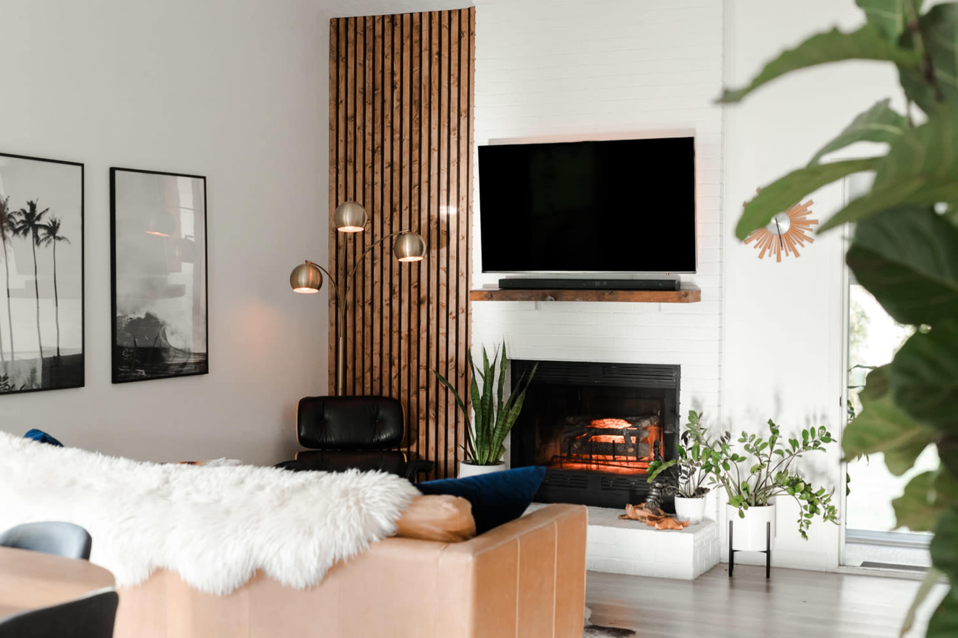 The living room features a leather sectional couch, a mounted flat-screen TV, a fireplace, and decorative plants against a backdrop of wooden wall accents and framed photographs.