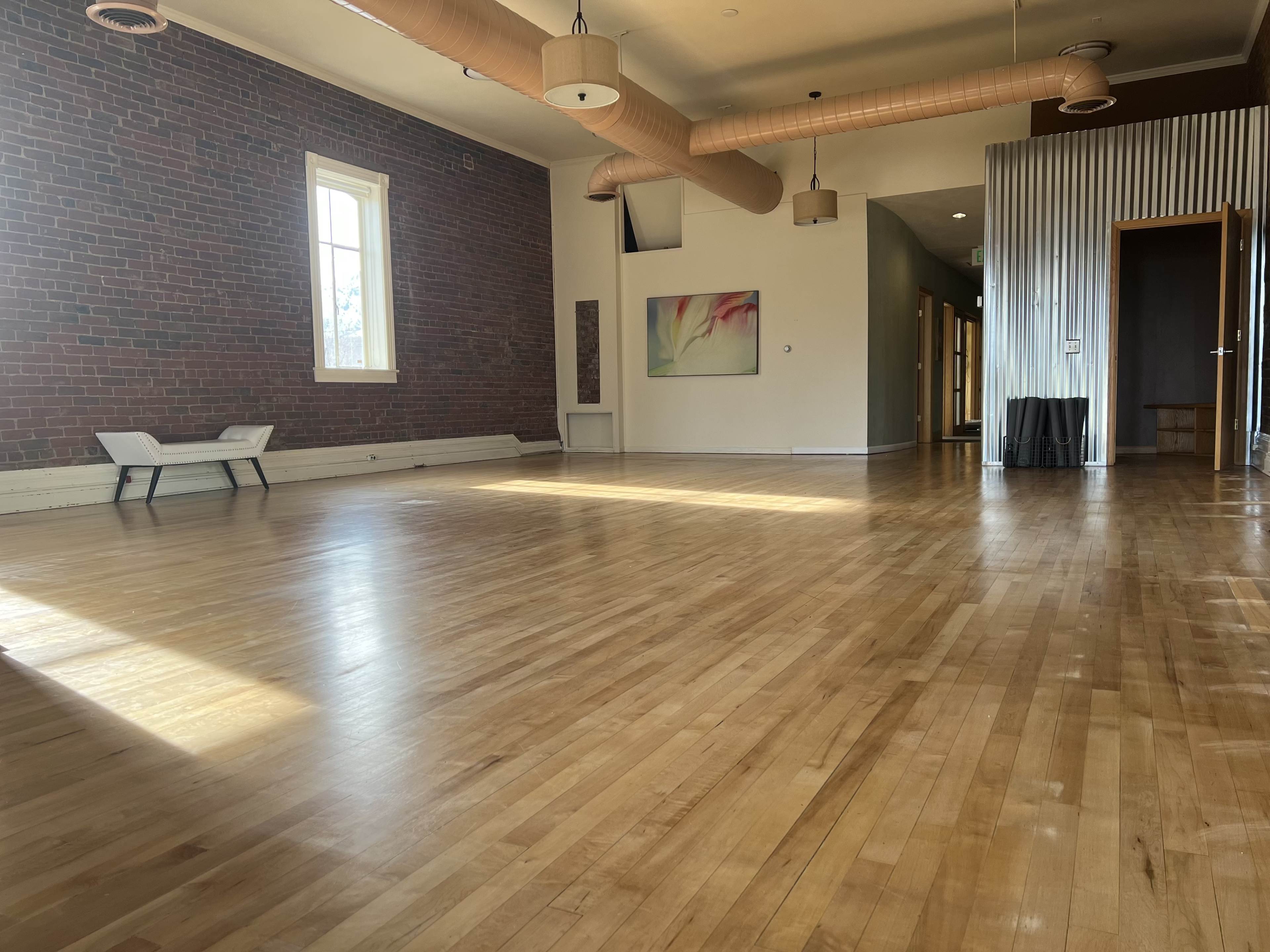 The image shows a spacious room with hardwood flooring, large windows, and a few pieces of furniture, including two chairs and a wall art piece.