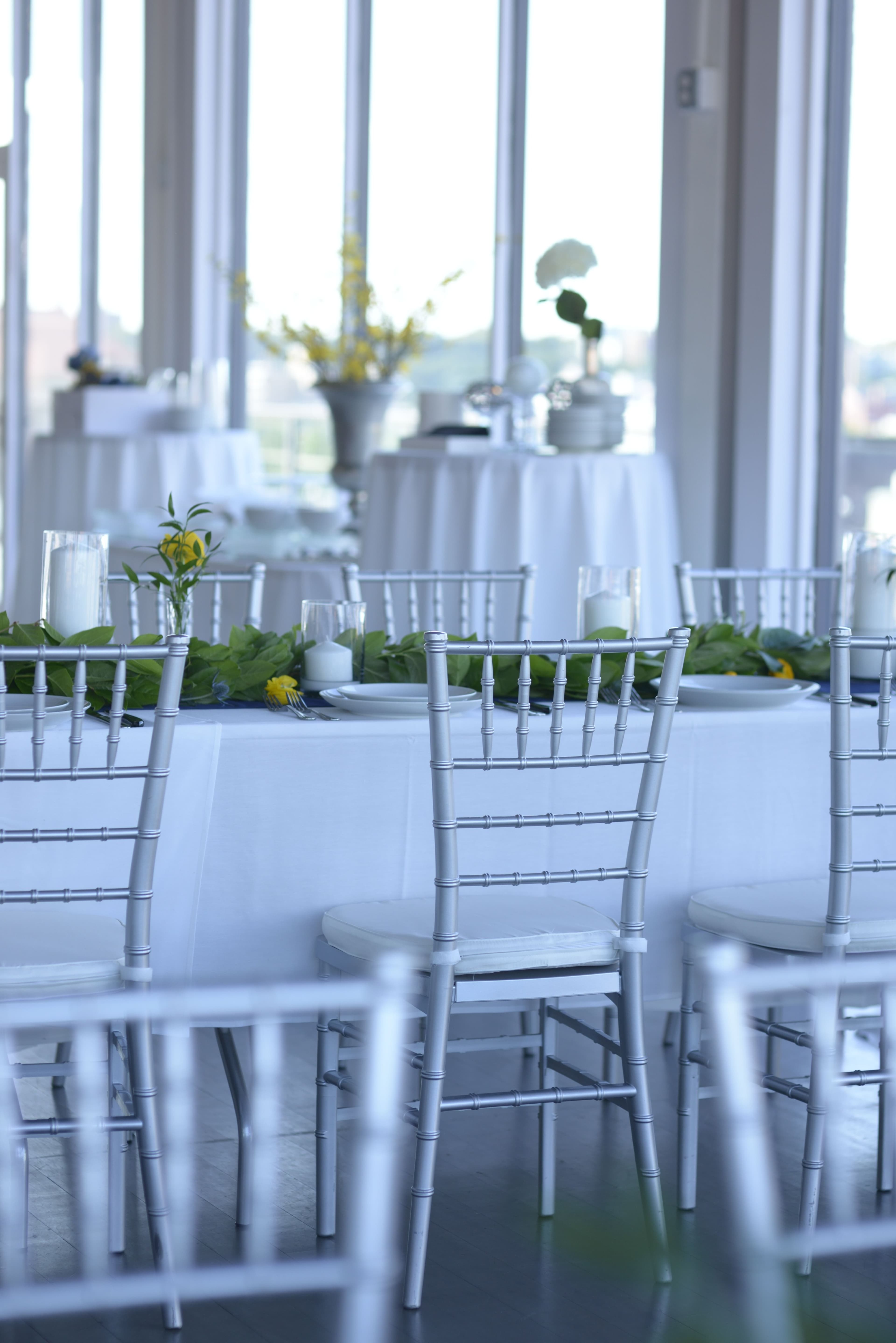 A spacious banquet room features tables set with white tablecloths and adorned with leafy centerpieces and drinks.
