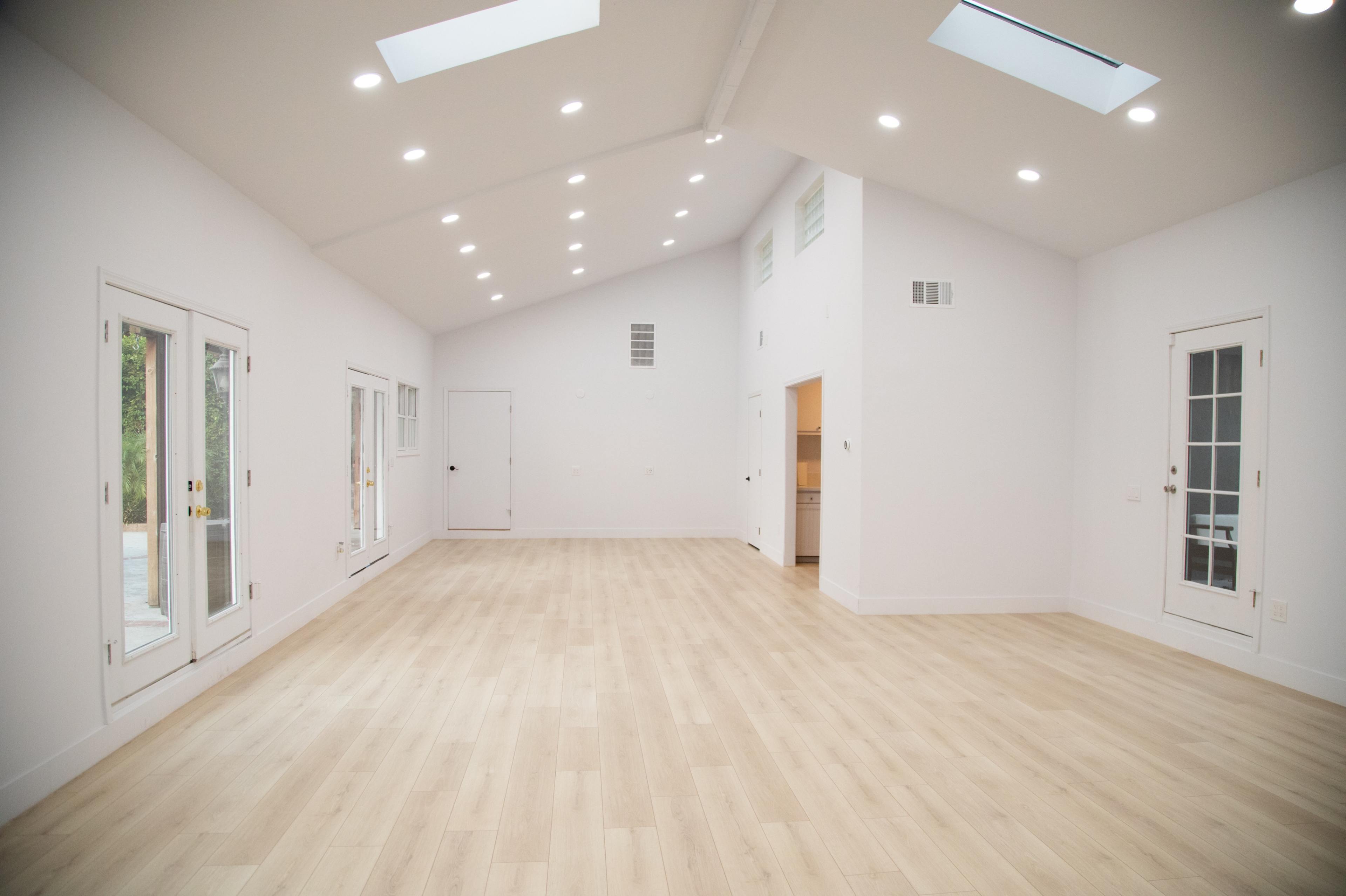 A spacious, empty room with white walls, wooden flooring, and multiple skylights and doors leading to an outdoor area.