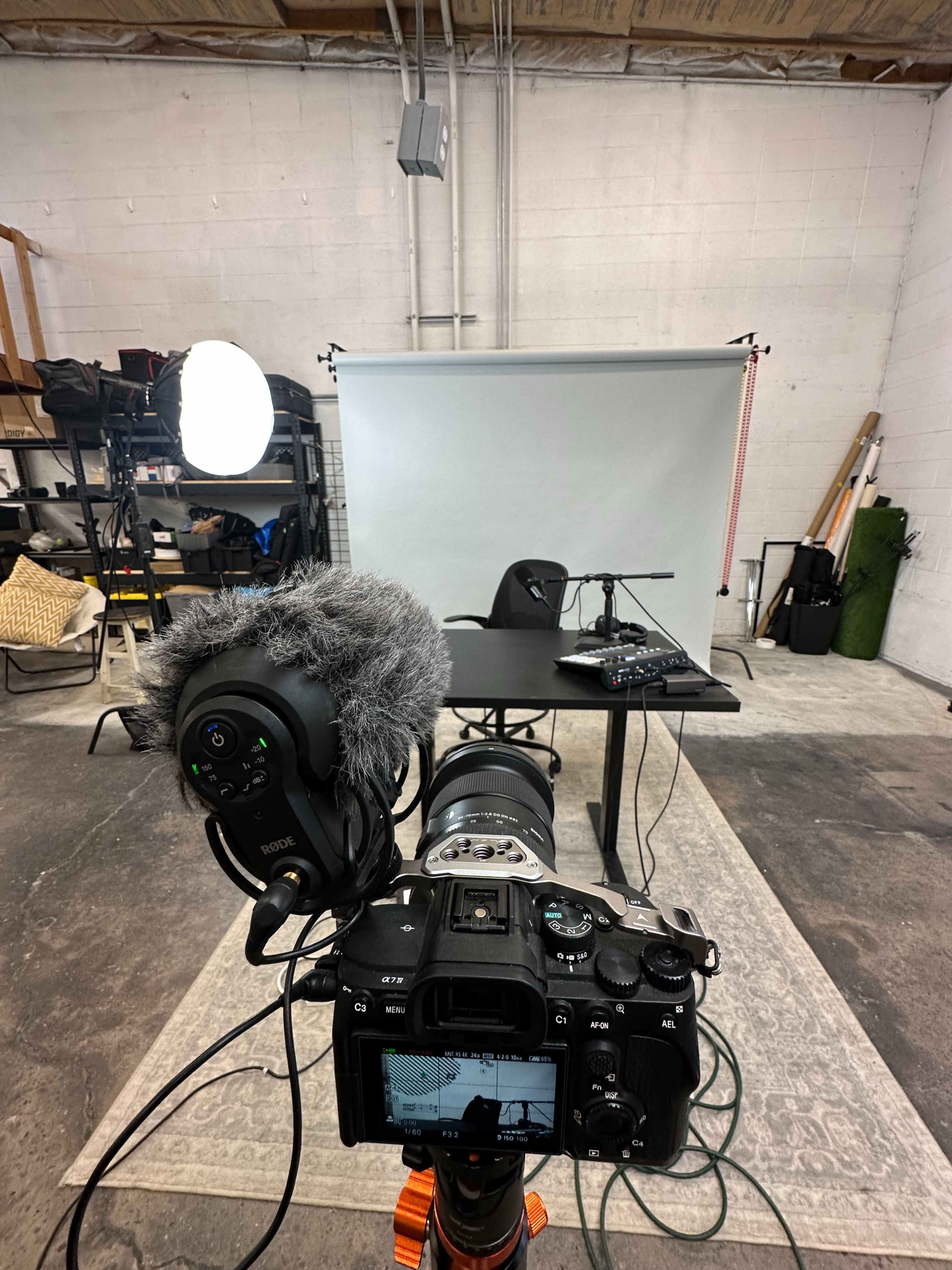 A camera is positioned on a tripod facing a table set up with a microphone and a laptop, surrounded by studio equipment and a plain backdrop in a well-lit room.