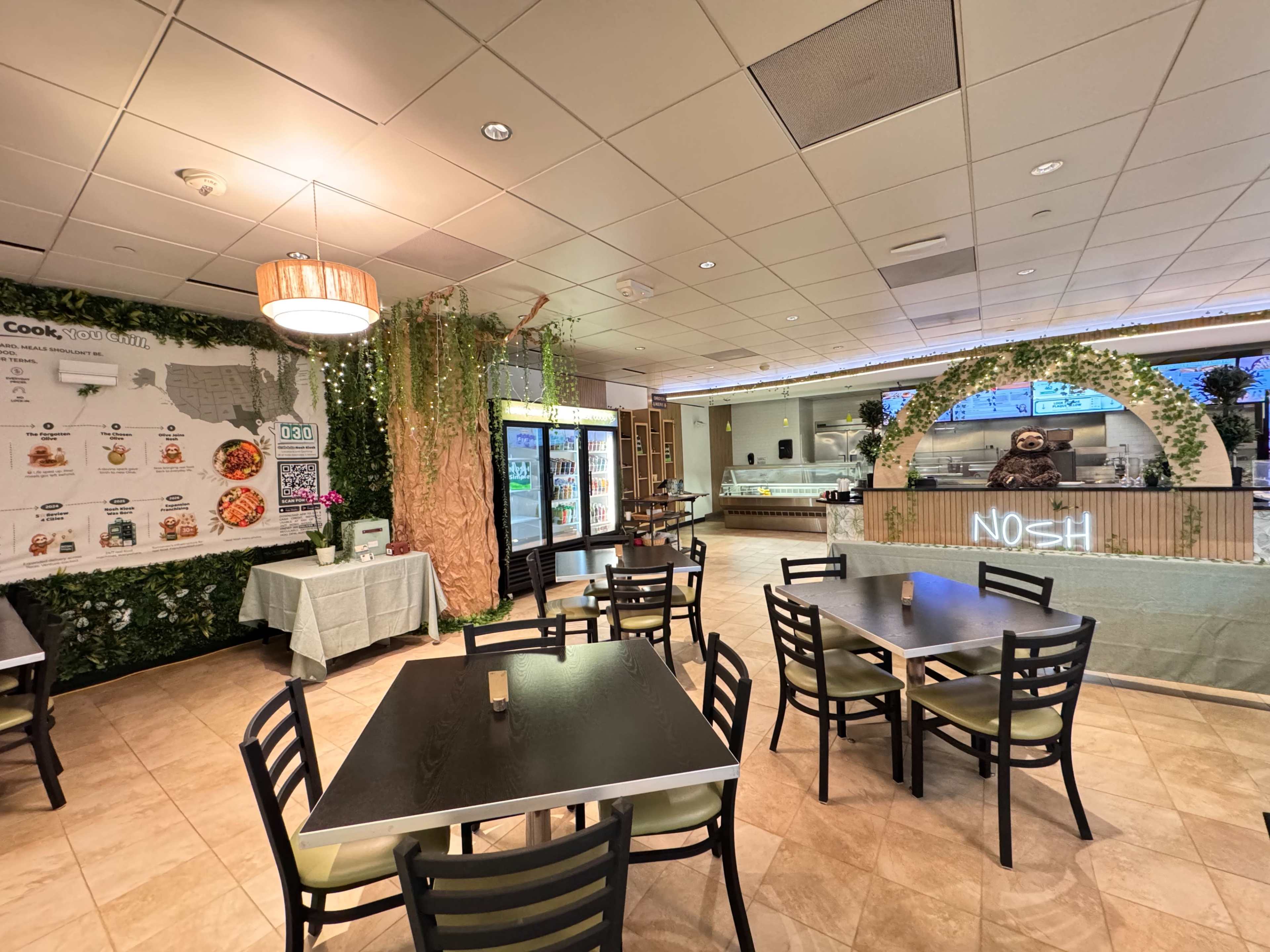 The image shows a modern dining area featuring several tables, a menu on the wall, a decorative tree, and a counter labeled "NOSH" with a glass display behind it.
