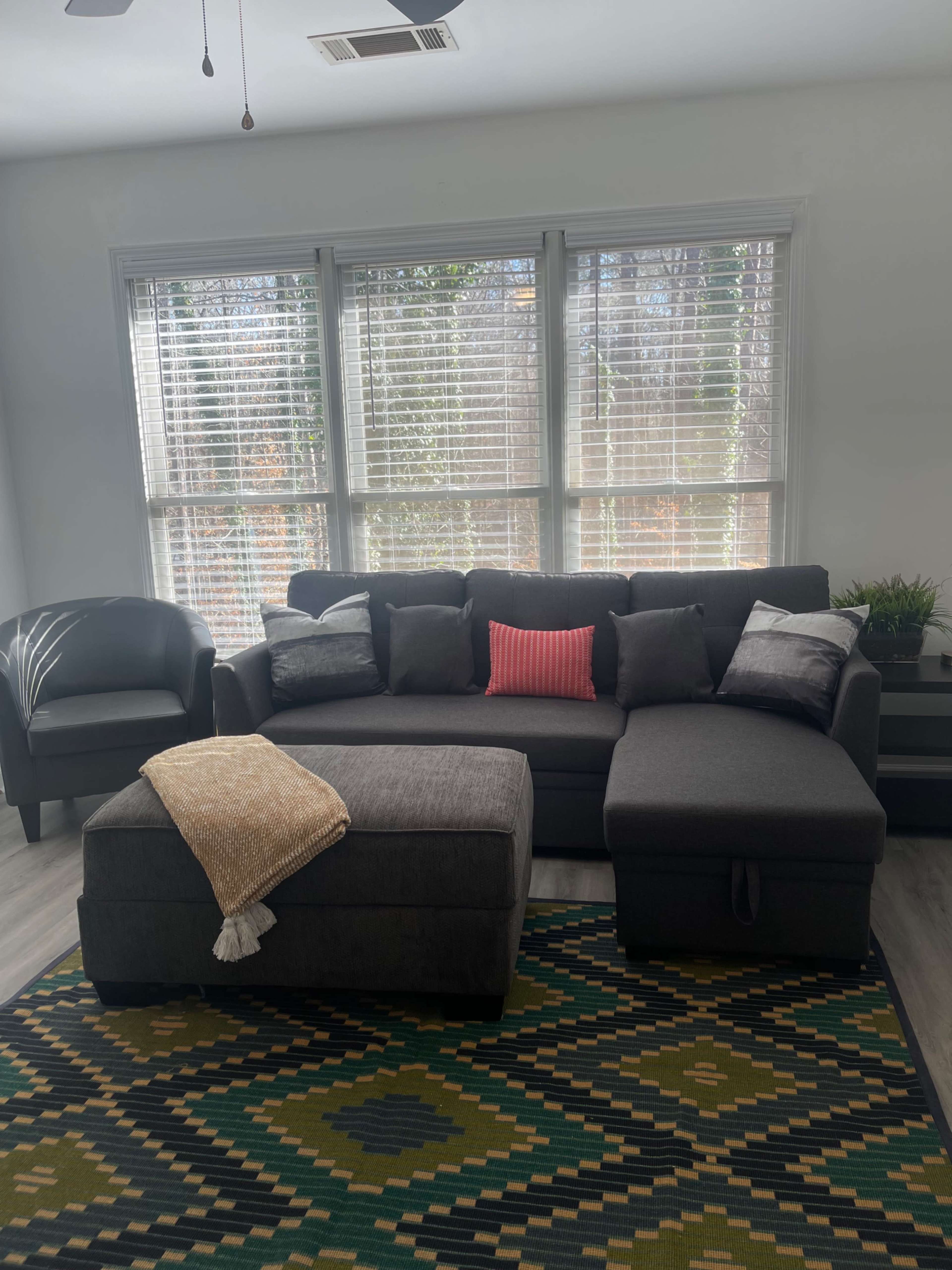 The image shows a living room featuring a dark gray sectional couch with pillows, an ottoman, a green and yellow patterned rug, and large windows allowing natural light to enter.