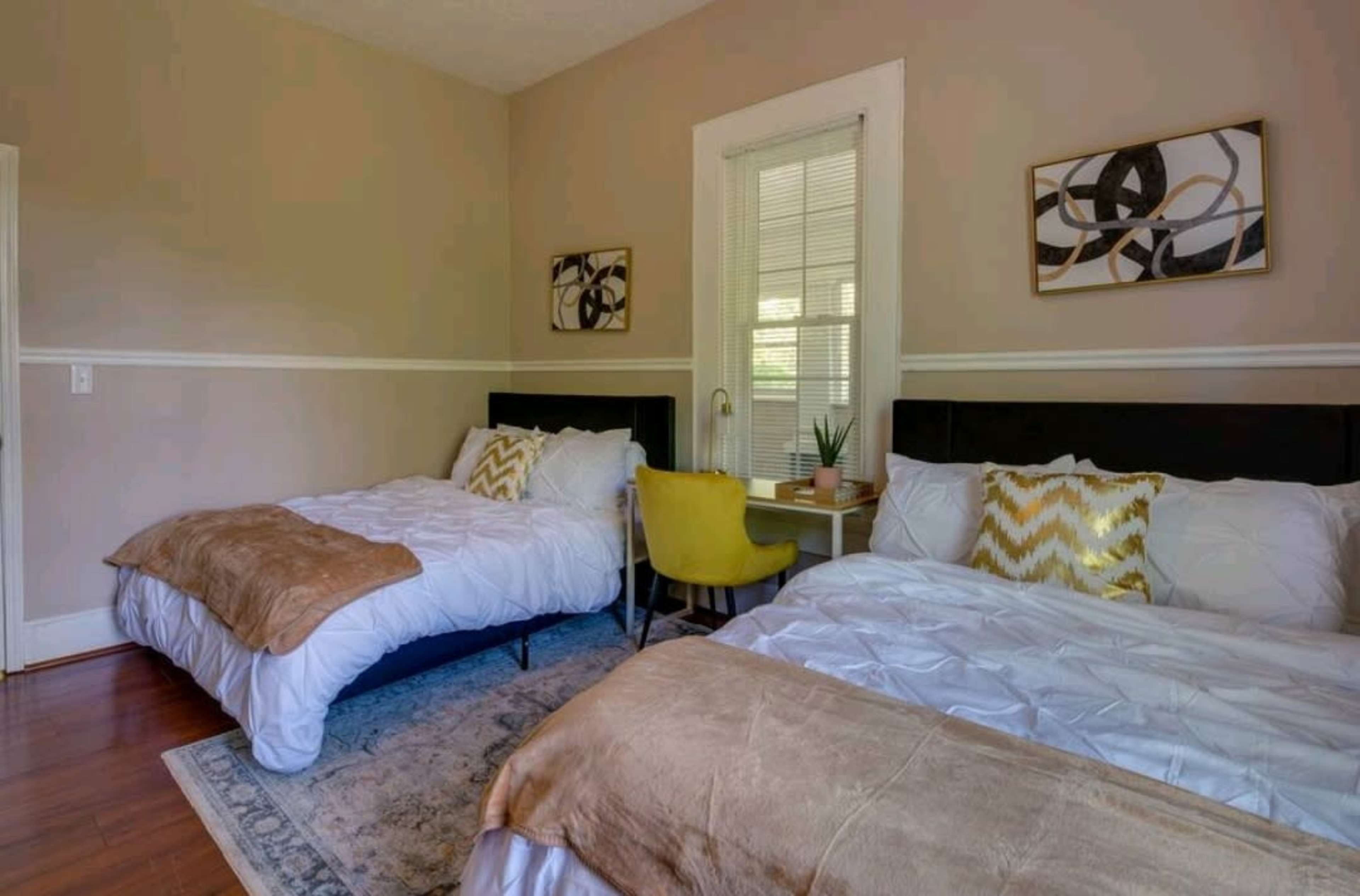 The image shows a cozy bedroom with two beds, each dressed in white bedding, a yellow accent chair, and framed wall art.