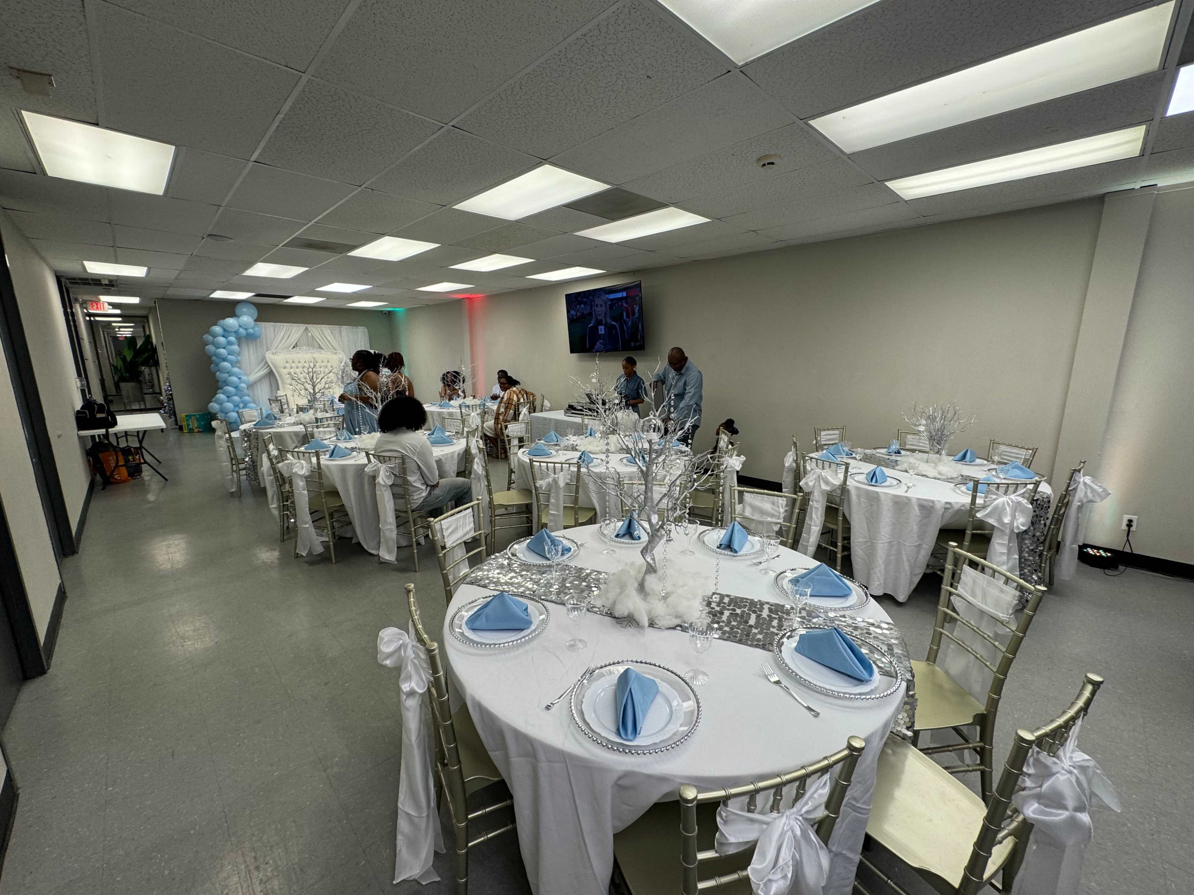 A banquet hall is set up for an event, featuring round tables covered in white tablecloths with blue accents and decorated with elegant centerpieces.