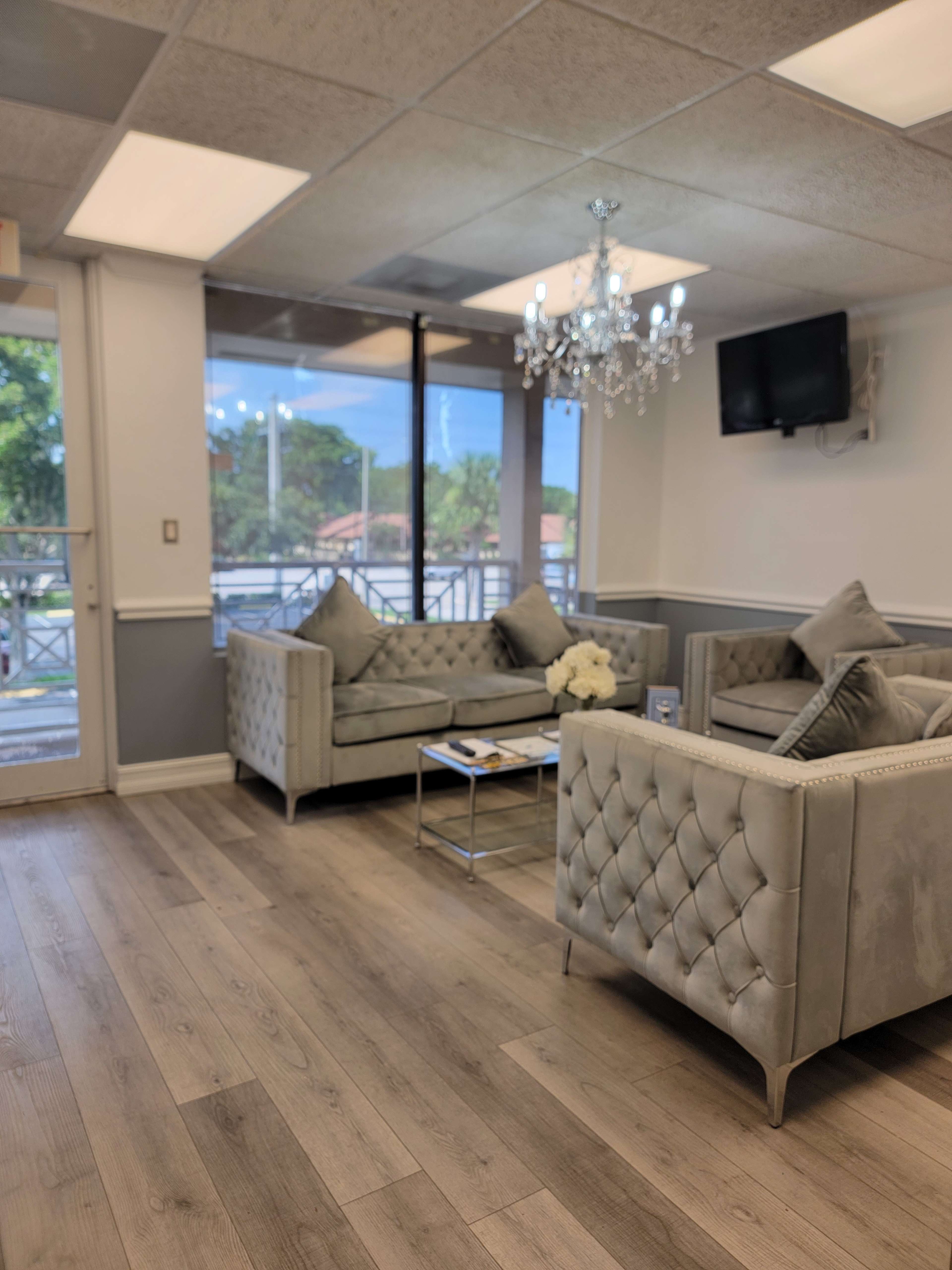 The image shows a modern waiting area featuring two light gray tufted sofas, a glass coffee table, and a chandelier, with large windows allowing natural light to enter.