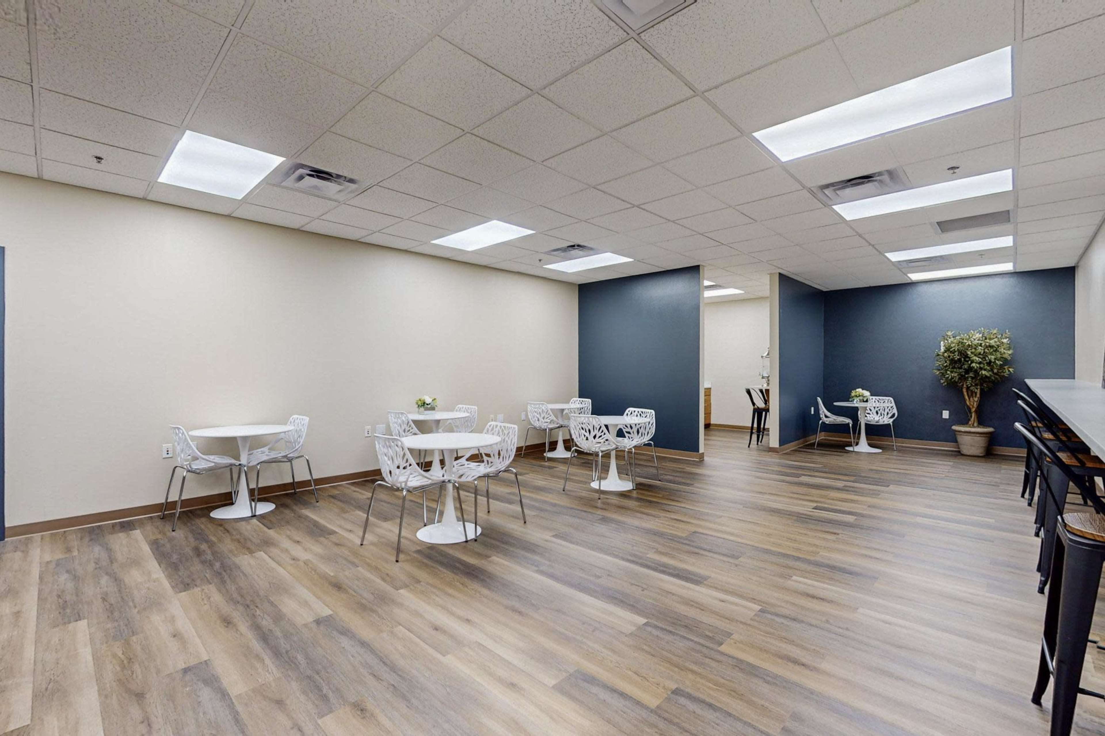 A modern, spacious room with multiple white tables and chairs, a wooden floor, and potted plants along the walls.