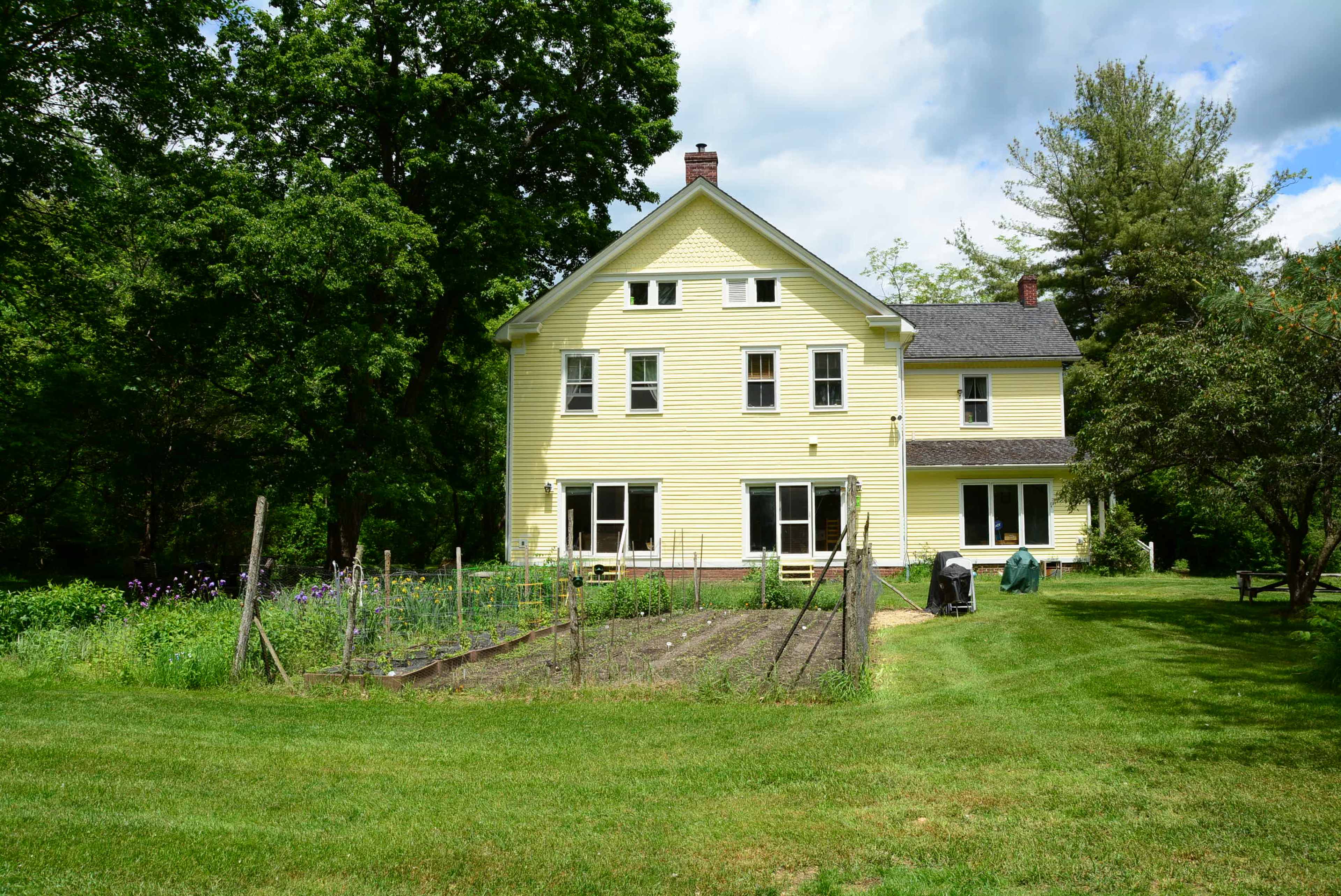 A two-story yellow house is situated behind a garden with rows of plants and a lawn surrounded by trees.