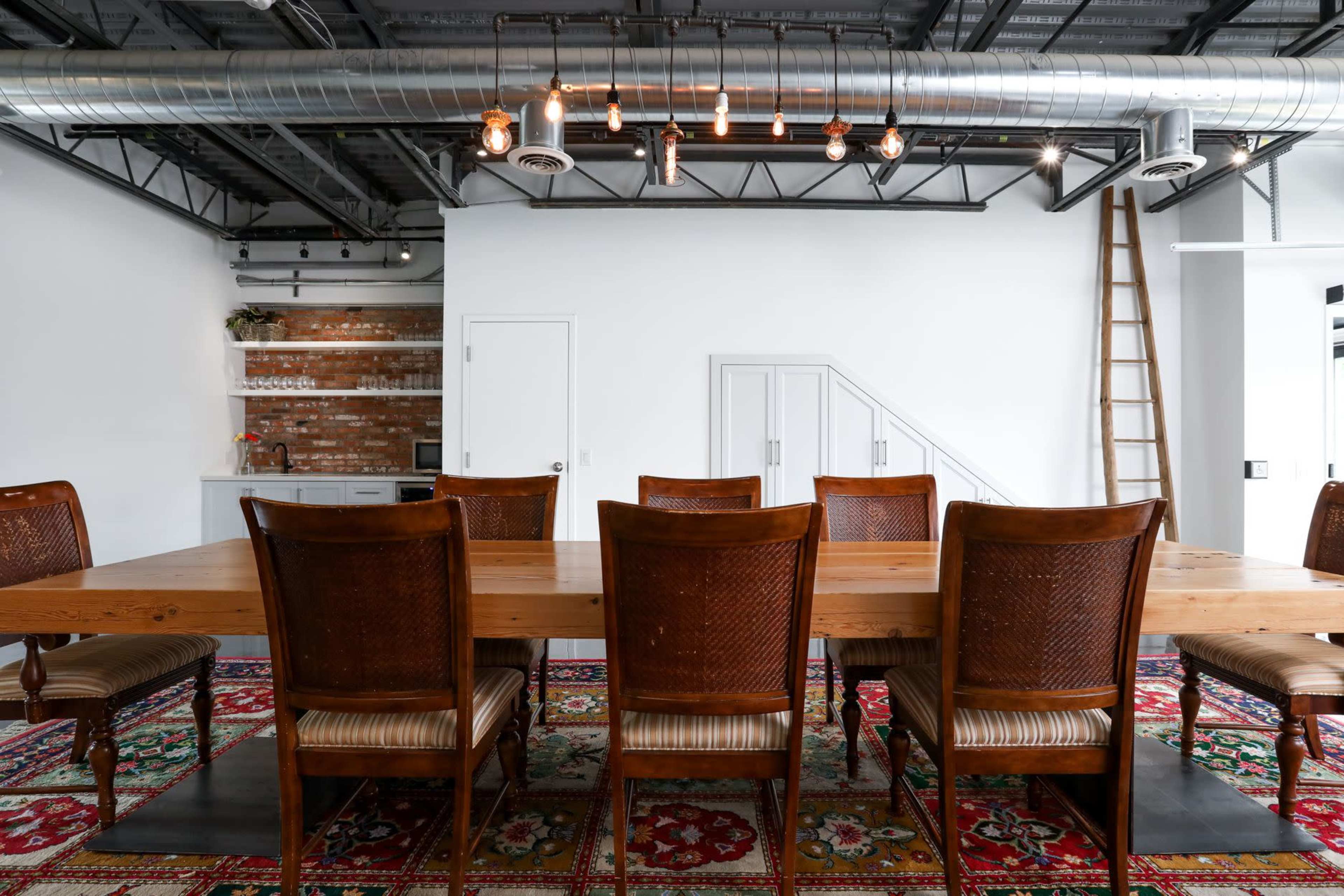 A long wooden dining table with eight brown chairs is set in a spacious room featuring exposed metal beams and soft lighting, with a patterned rug underneath.