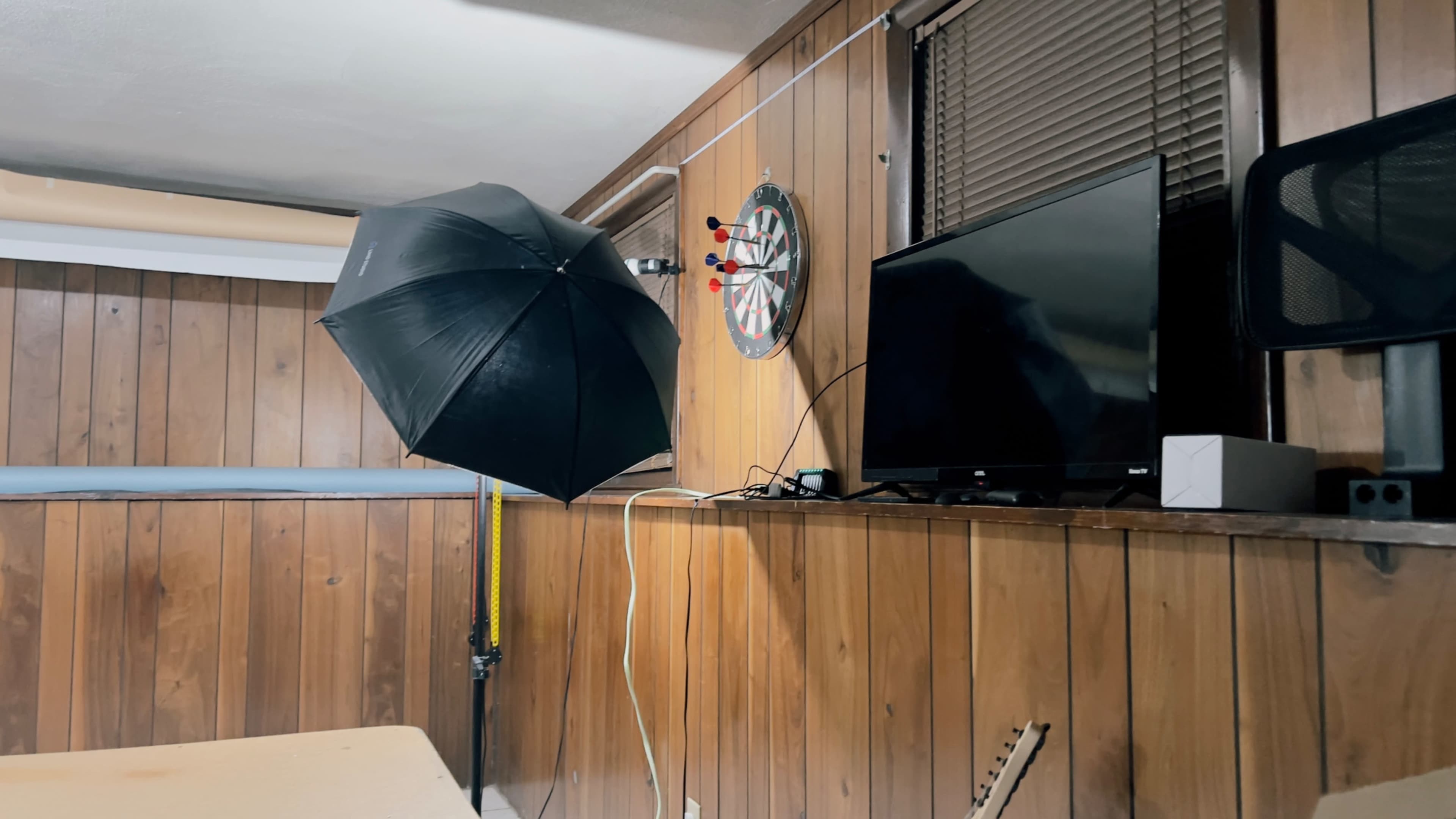 A wooden-paneled room featuring a black umbrella light, a dartboard on the wall, and a television mounted next to it.