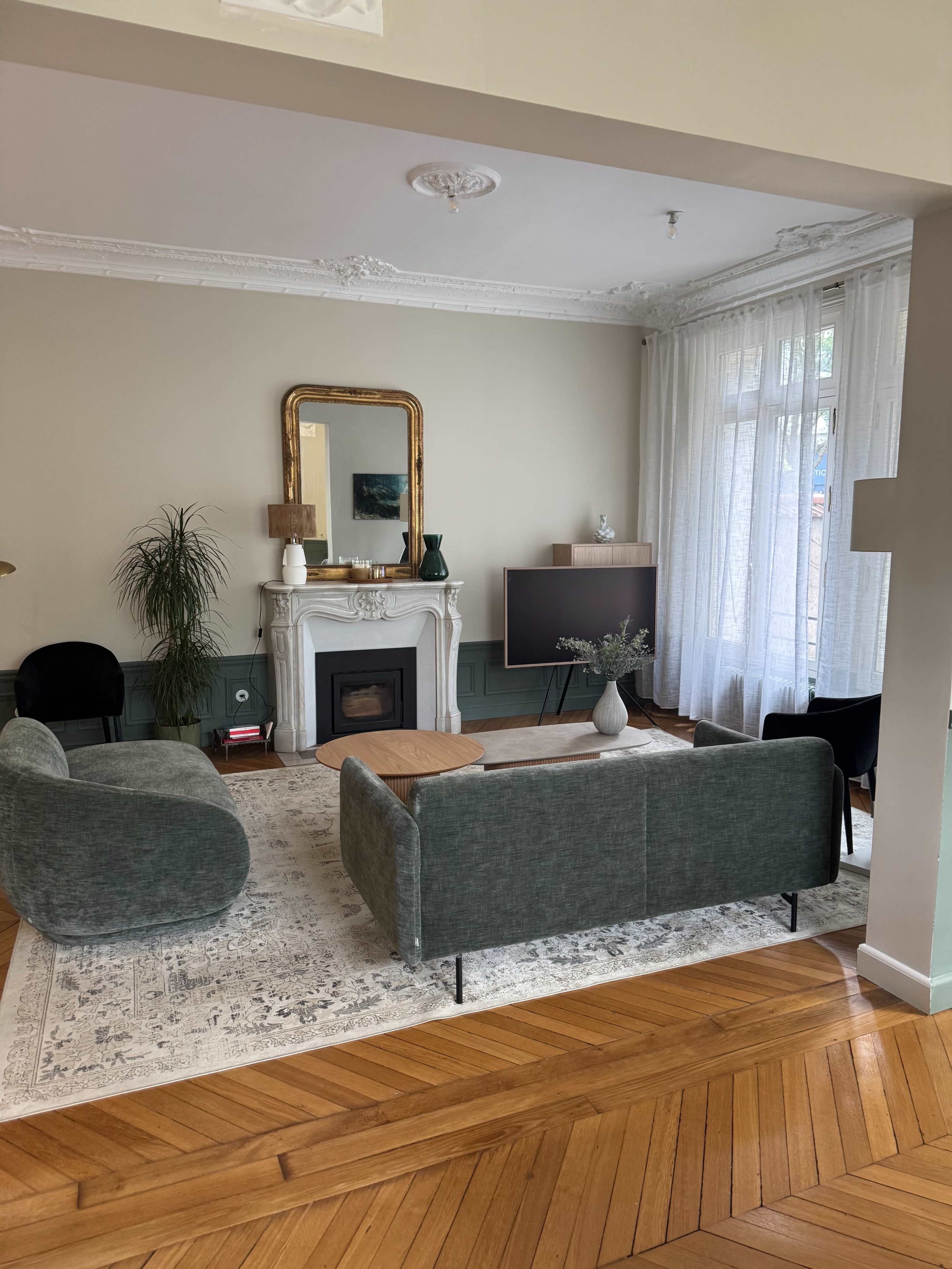 The living room features a pair of sofas facing a fireplace with a decorative mirror above it, surrounded by plants and natural light filtering through sheer curtains.