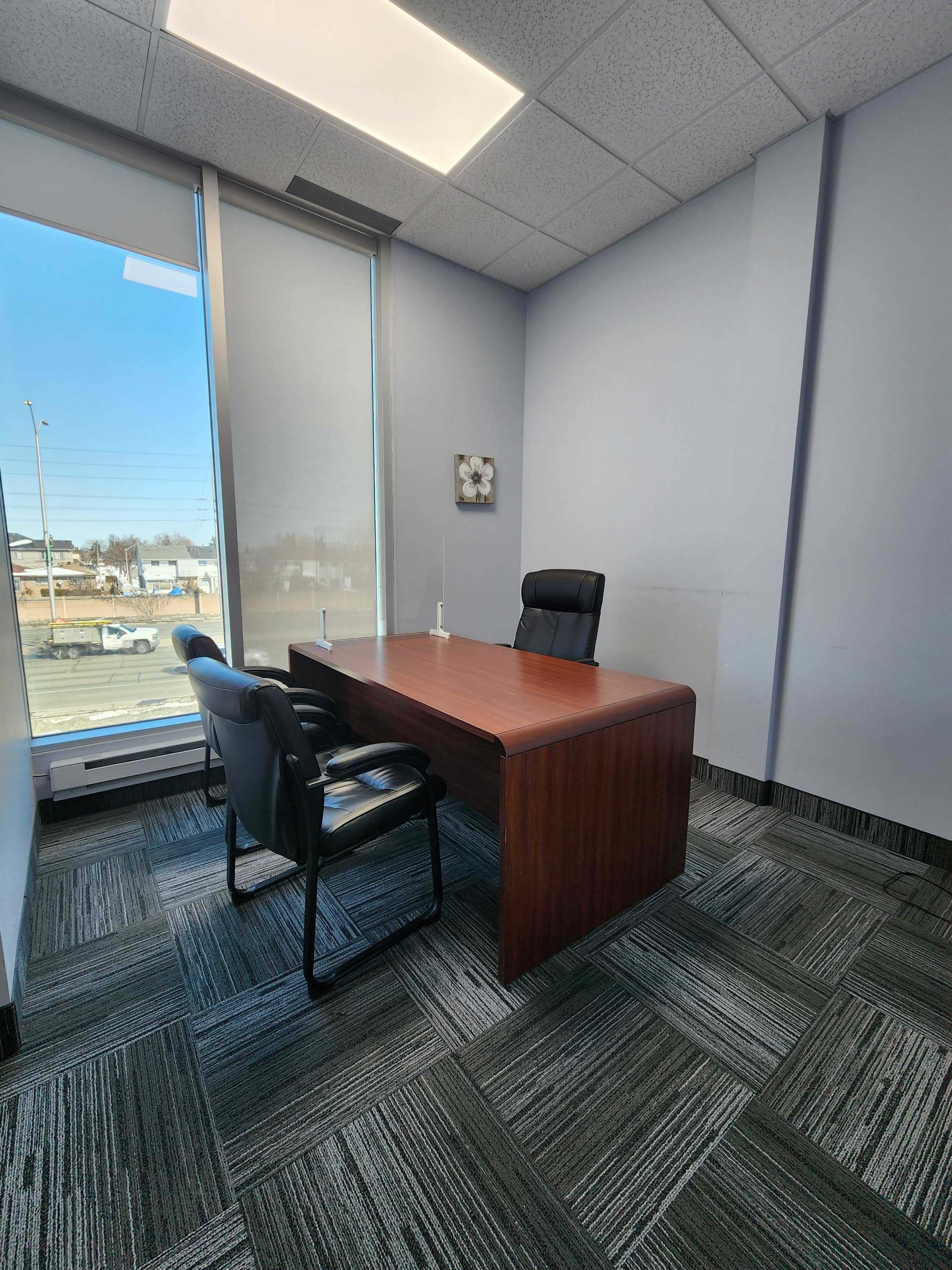A modern office with a wooden desk and two black chairs, featuring large windows that allow natural light to enter the space.