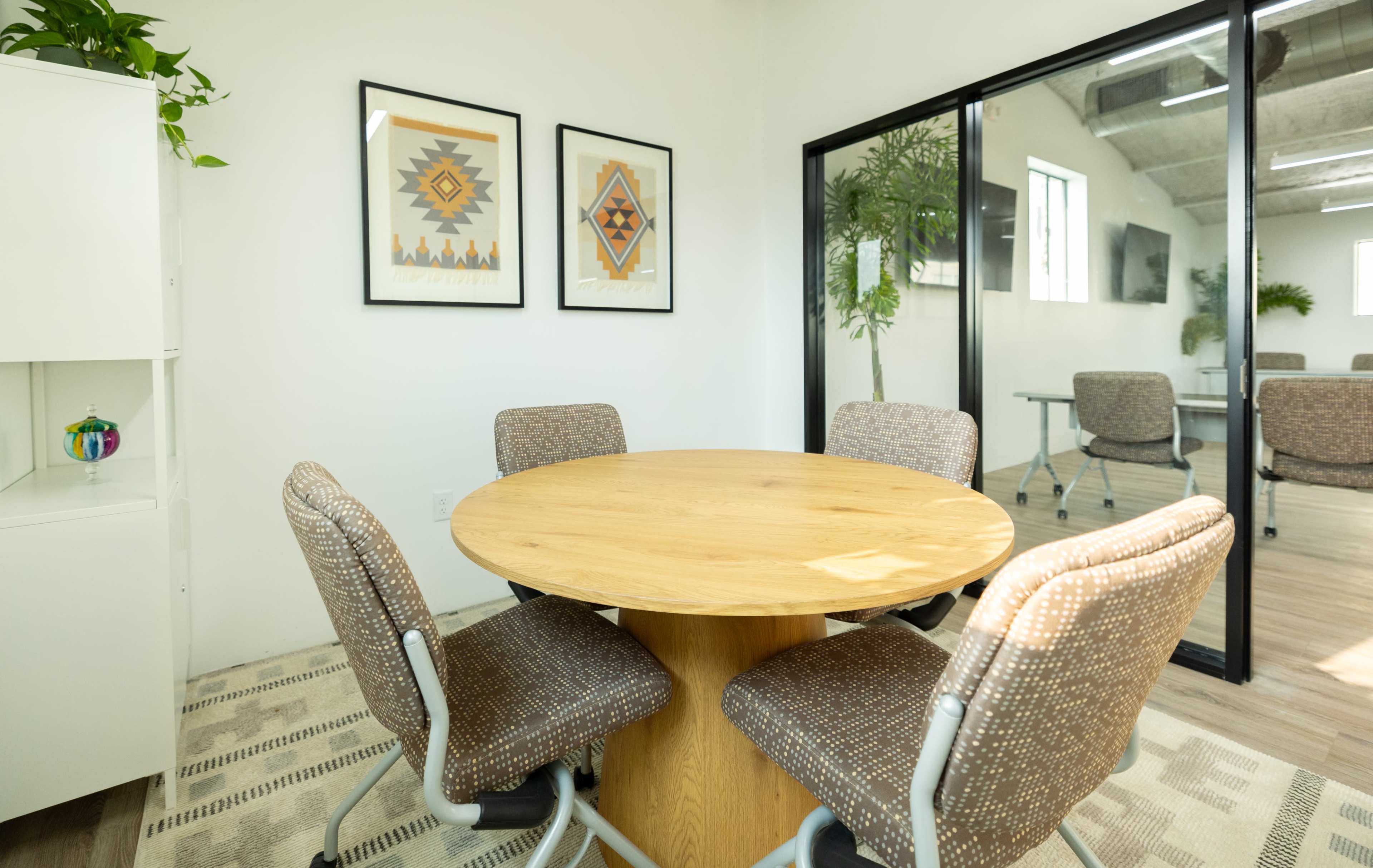A small conference room featuring a round wooden table surrounded by four patterned chairs, with framed artwork on the walls and glass partitions.