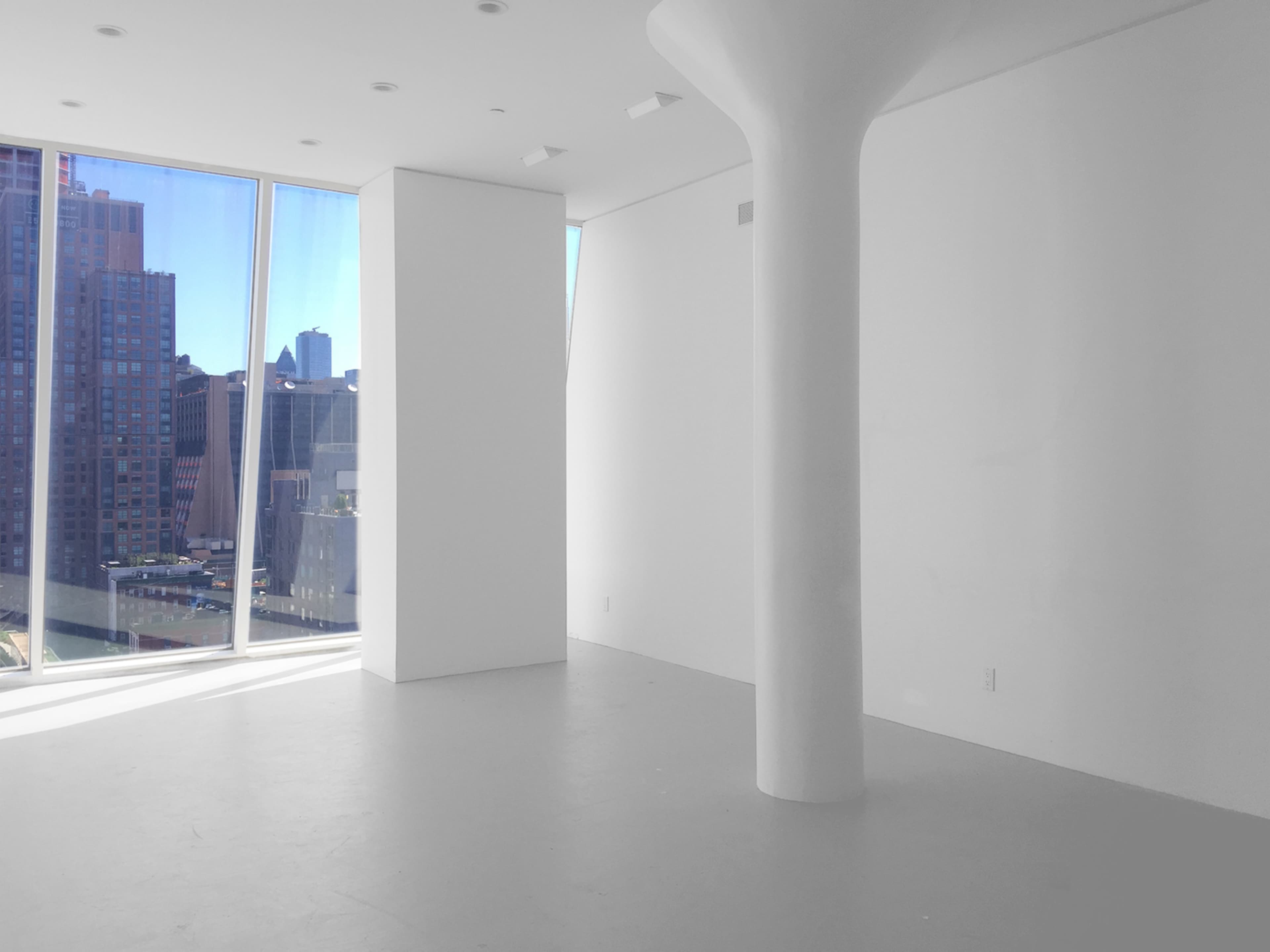 An empty, minimalist room features large windows with city views and a central white column.