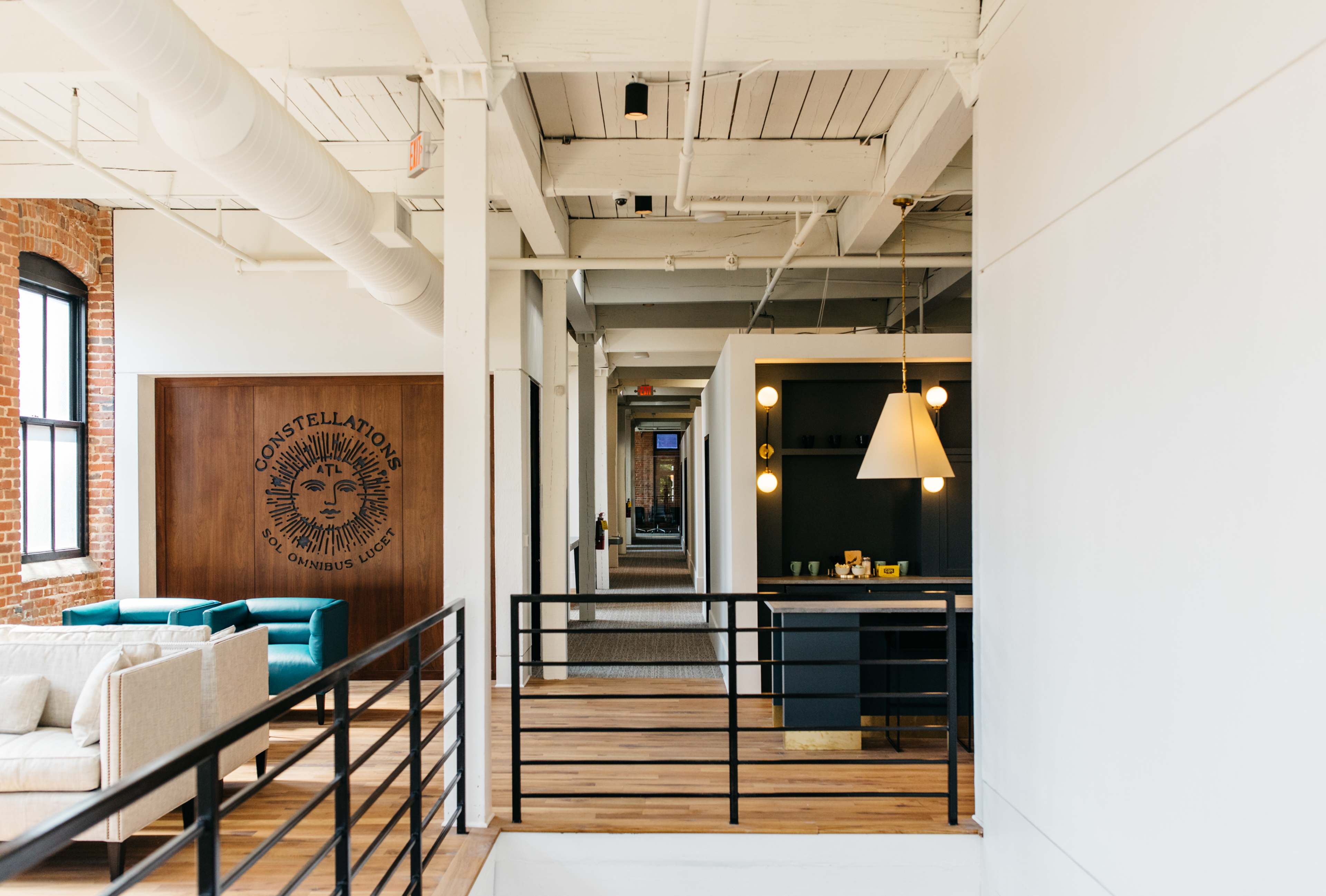 The image shows a modern office interior with a long corridor featuring a lounge area, wooden flooring, and a decorative wall with the logo "Constellations."