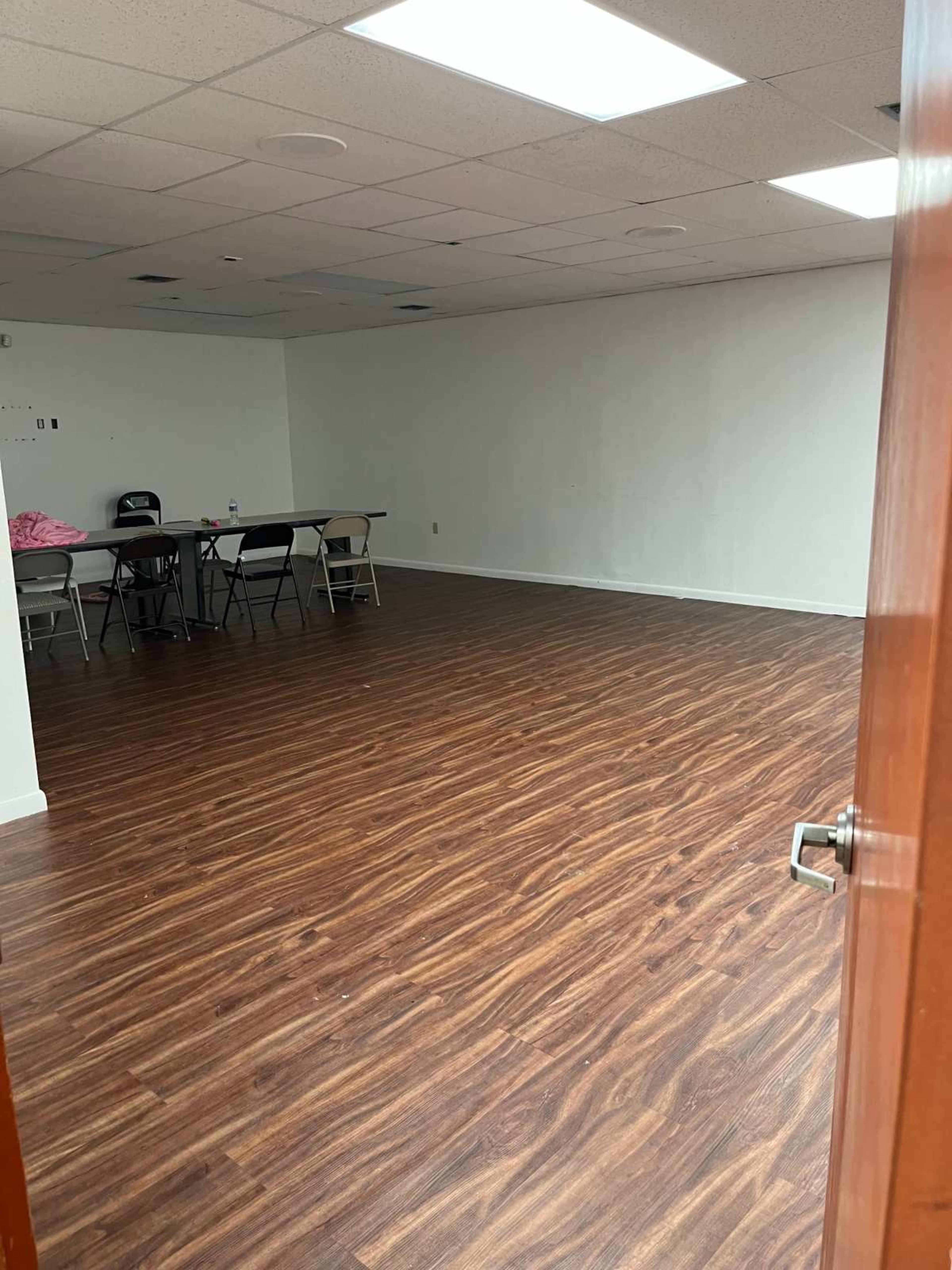 The image shows an empty room with wooden floors, a white wall, and a few folding chairs arranged around a table.