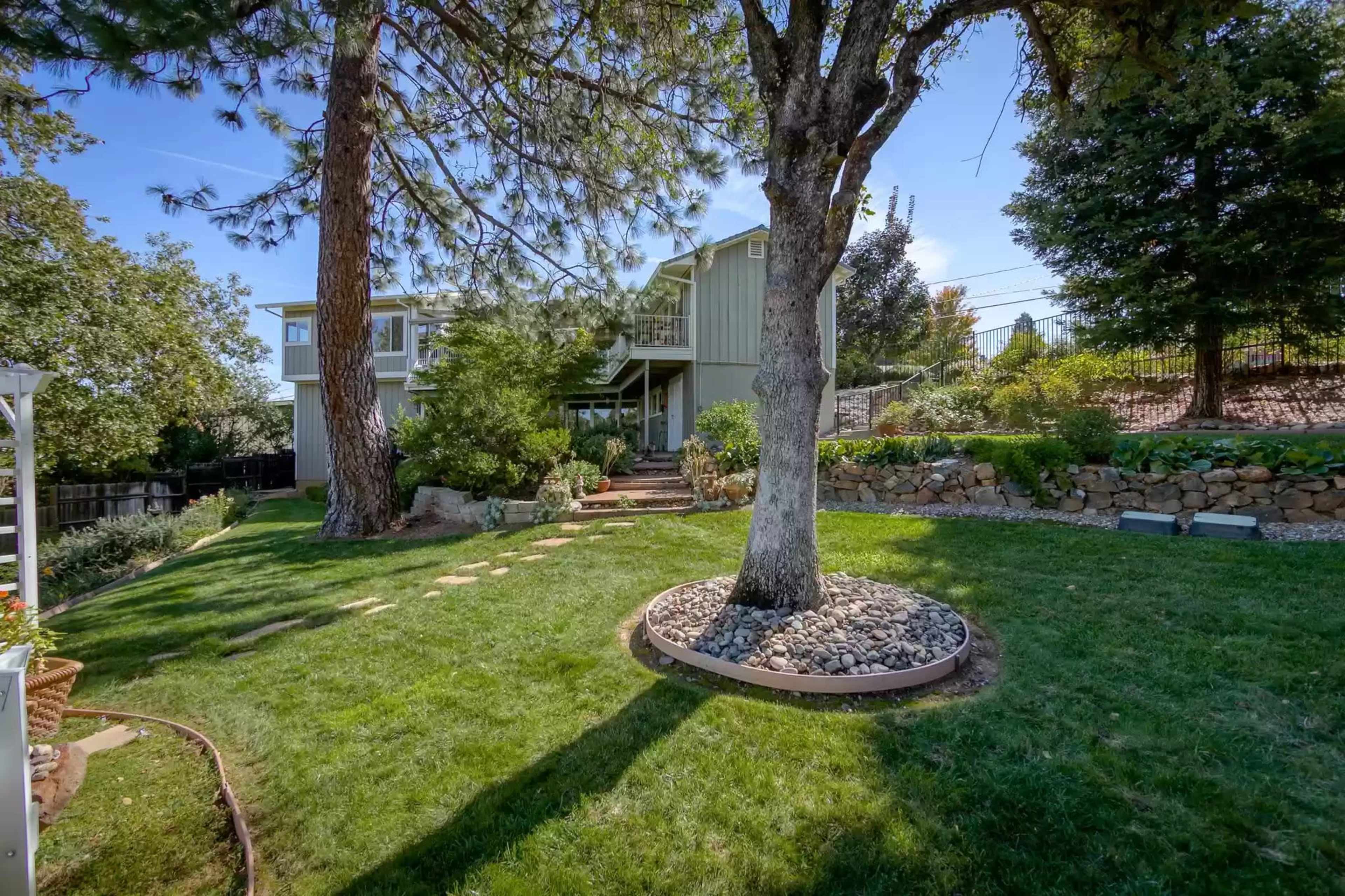 A two-story house surrounded by a landscaped yard featuring a large tree, stone pathways, and greenery.