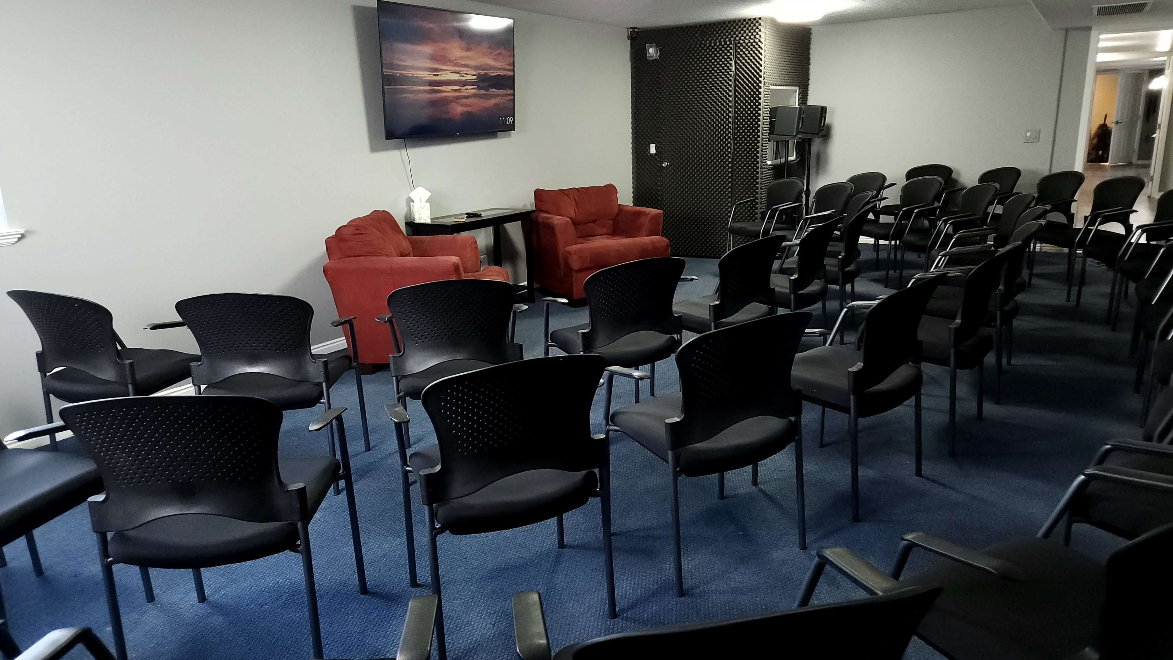 A room is set up for a meeting, featuring two red chairs with a table and a television mounted on the wall, surrounded by rows of black chairs on a blue carpet.