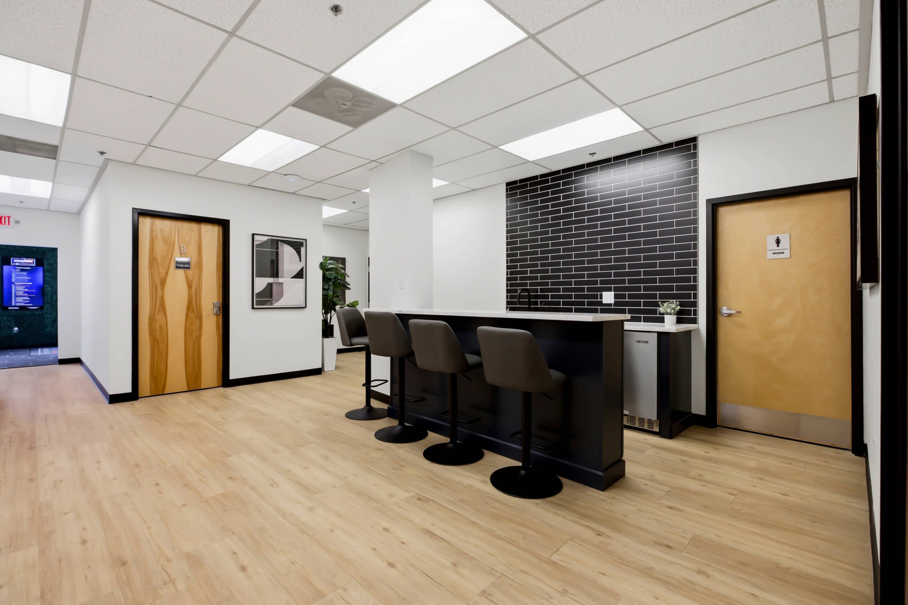 The image shows a modern office kitchenette area with a bar counter and stools, adjacent to a hallway featuring two doors.